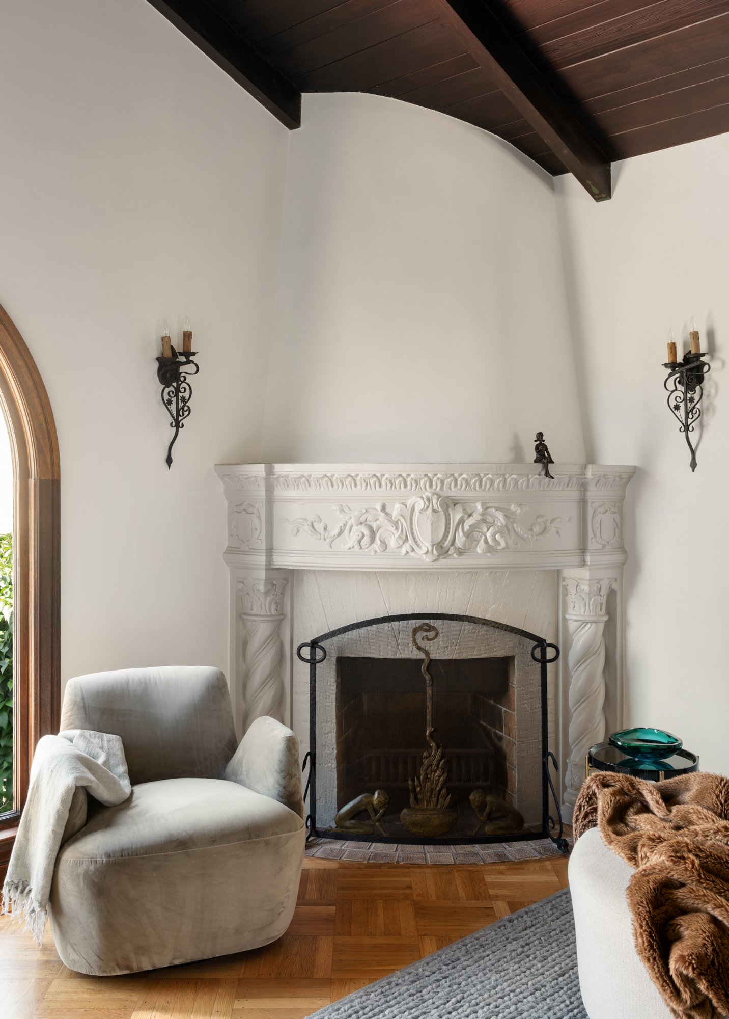 Living room with a white ornate fireplace, beige armchair with a white throw, a second beige armchair with a brown fur throw, a black metal fireplace screen, wall sconces, and hardwood floors.