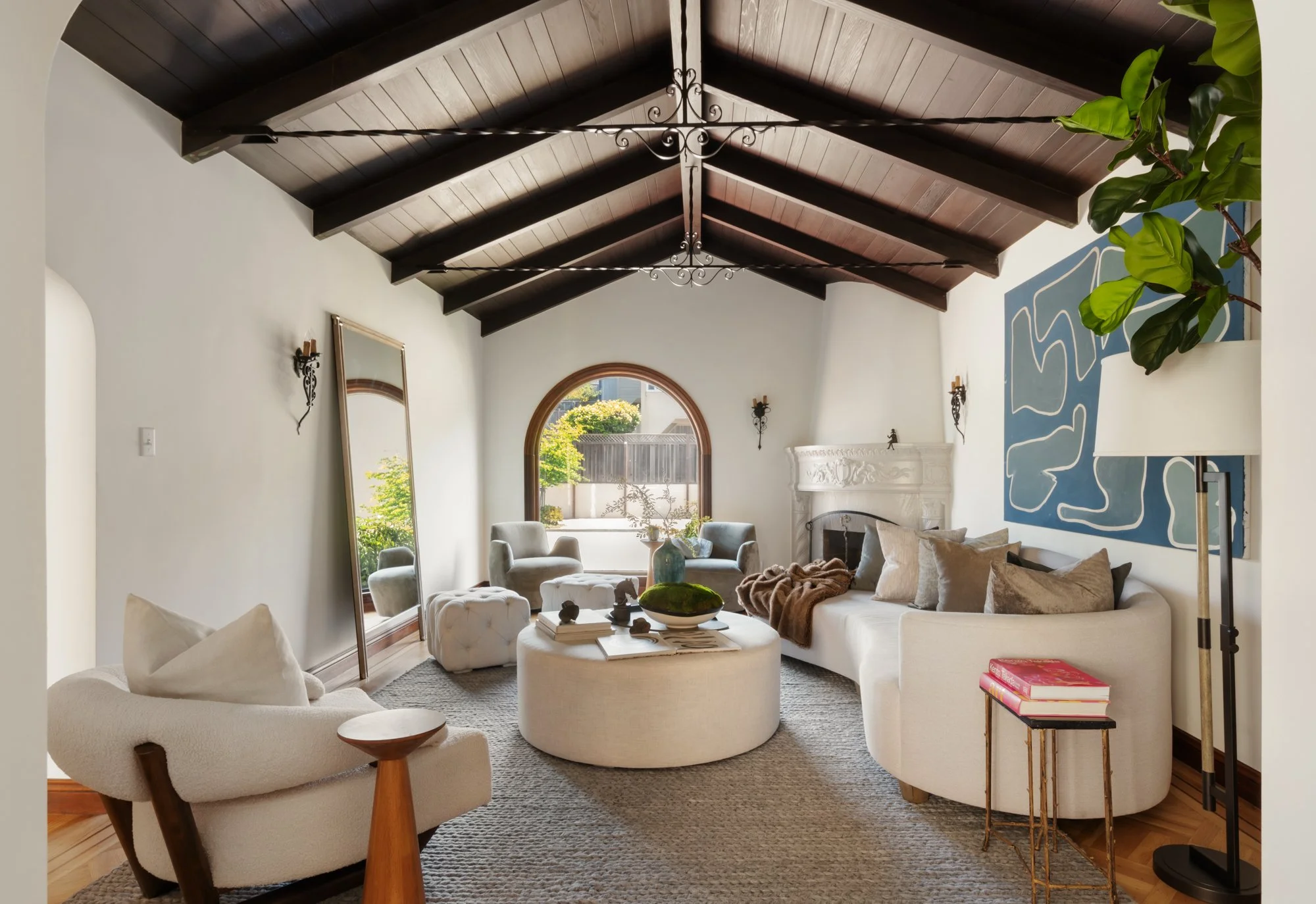 Living room with white couches, a round ottoman, a large abstract artwork on the wall, and a fireplace. The ceiling has dark wooden beams and wrought iron chandelier. Large window with outdoor greenery visible.