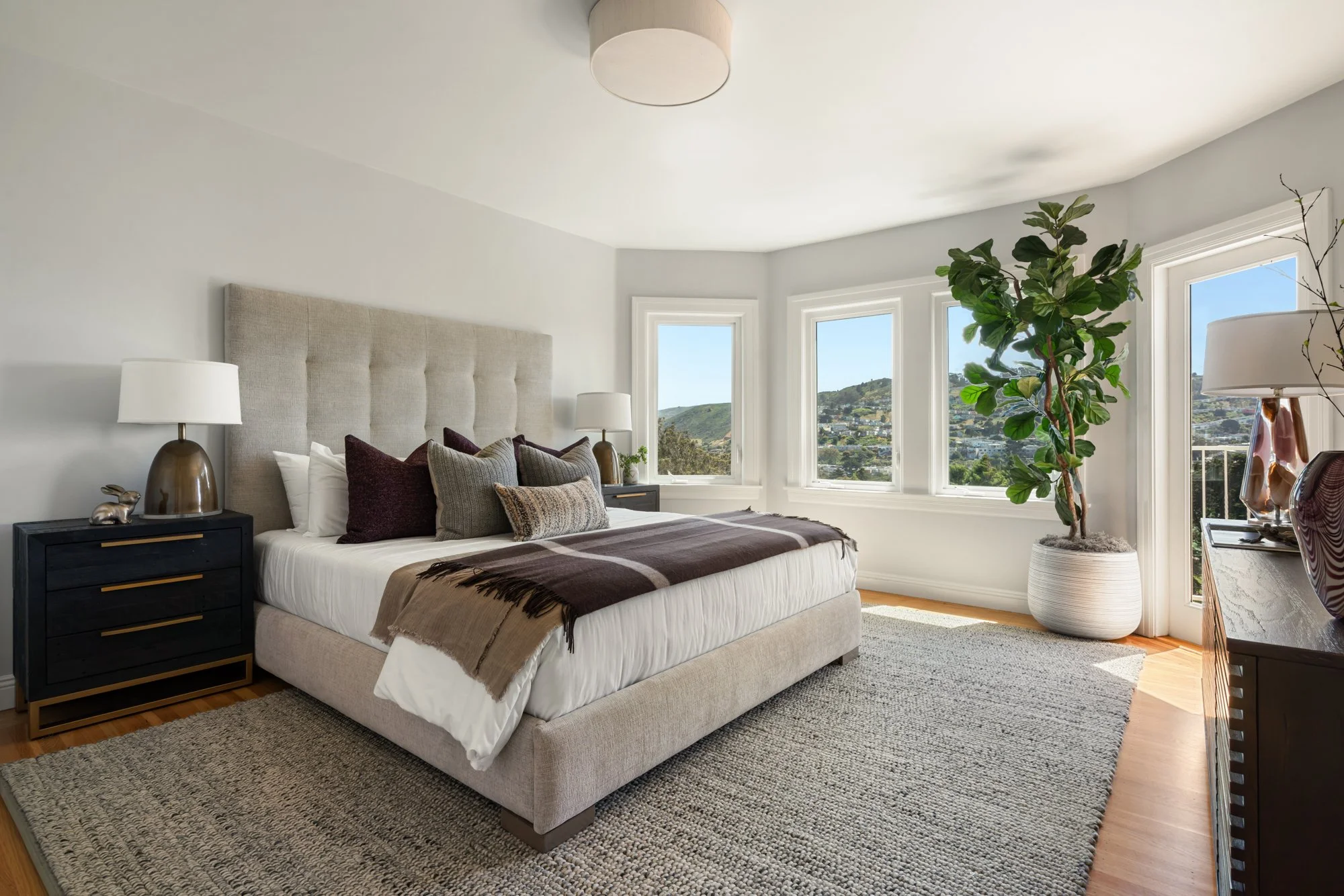 A bedroom with a beige upholstered bed, dark nightstands, white lamps, and a large potted fiddle leaf fig tree near three windows with city and hillside views. The room has white walls, a beige area rug, and light hardwood floors.