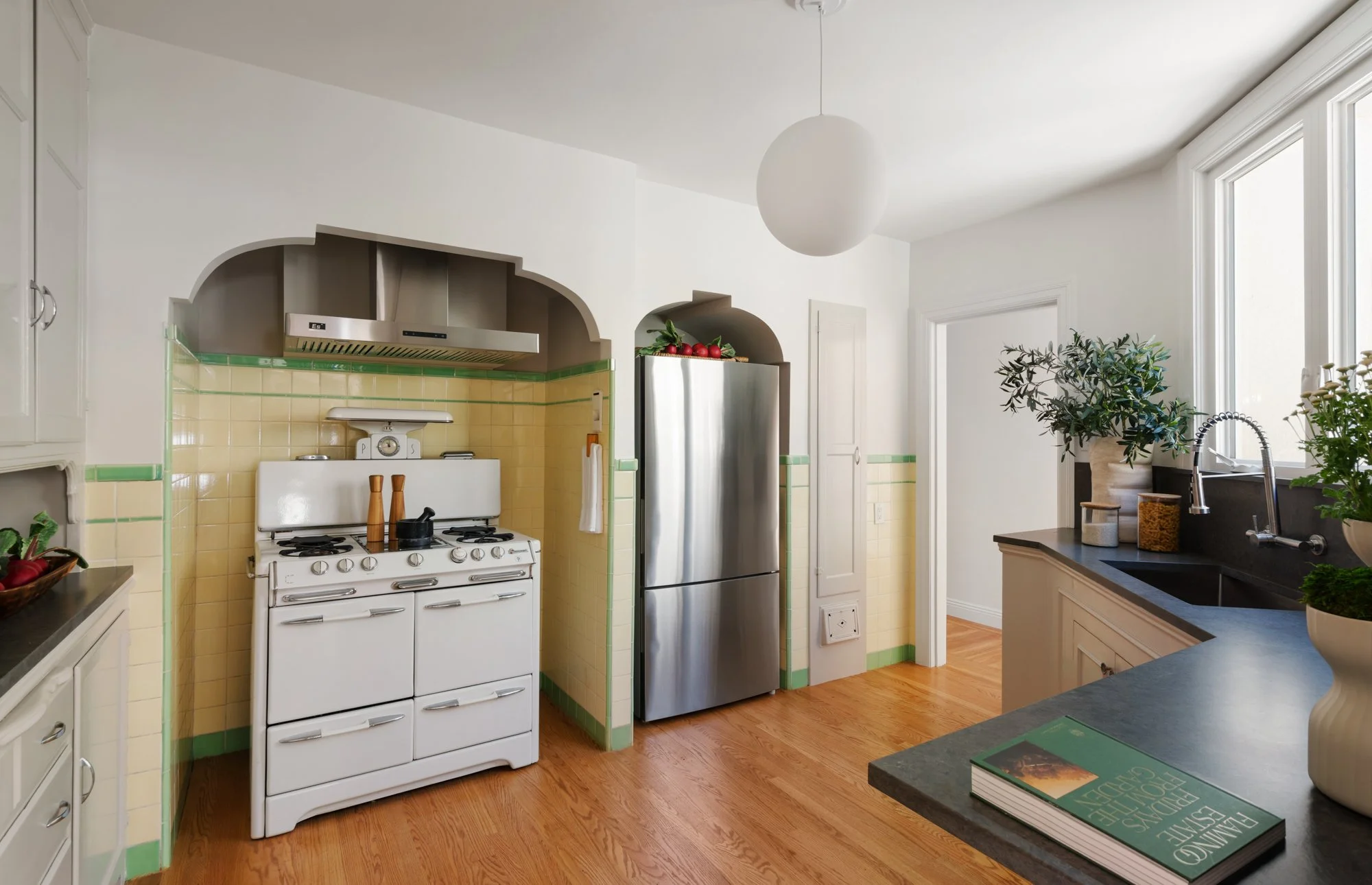 Kitchen with white stove, stainless steel refrigerator, black countertop, potted plants, and large windows.