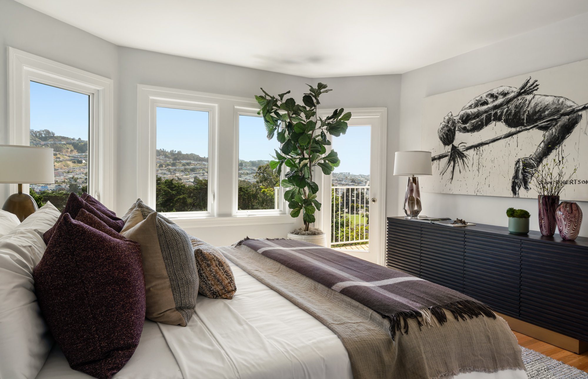 Living room with large windows, houseplants, a dark sideboard, abstract wall art, and a bed with purple and beige pillows.