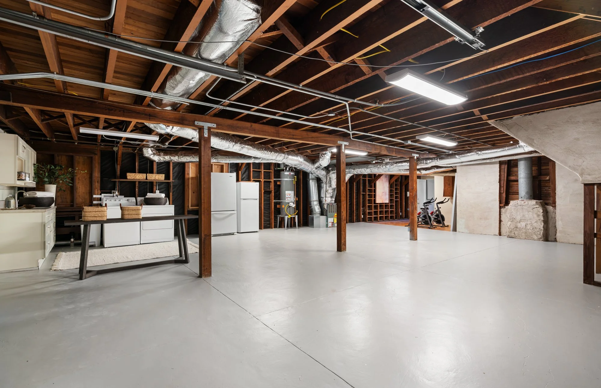 Empty basement or garage with unfinished wooden ceiling, exposed ductwork, and plain concrete floor. Contains a small kitchenette with white appliances, a table, and two exercise bikes.