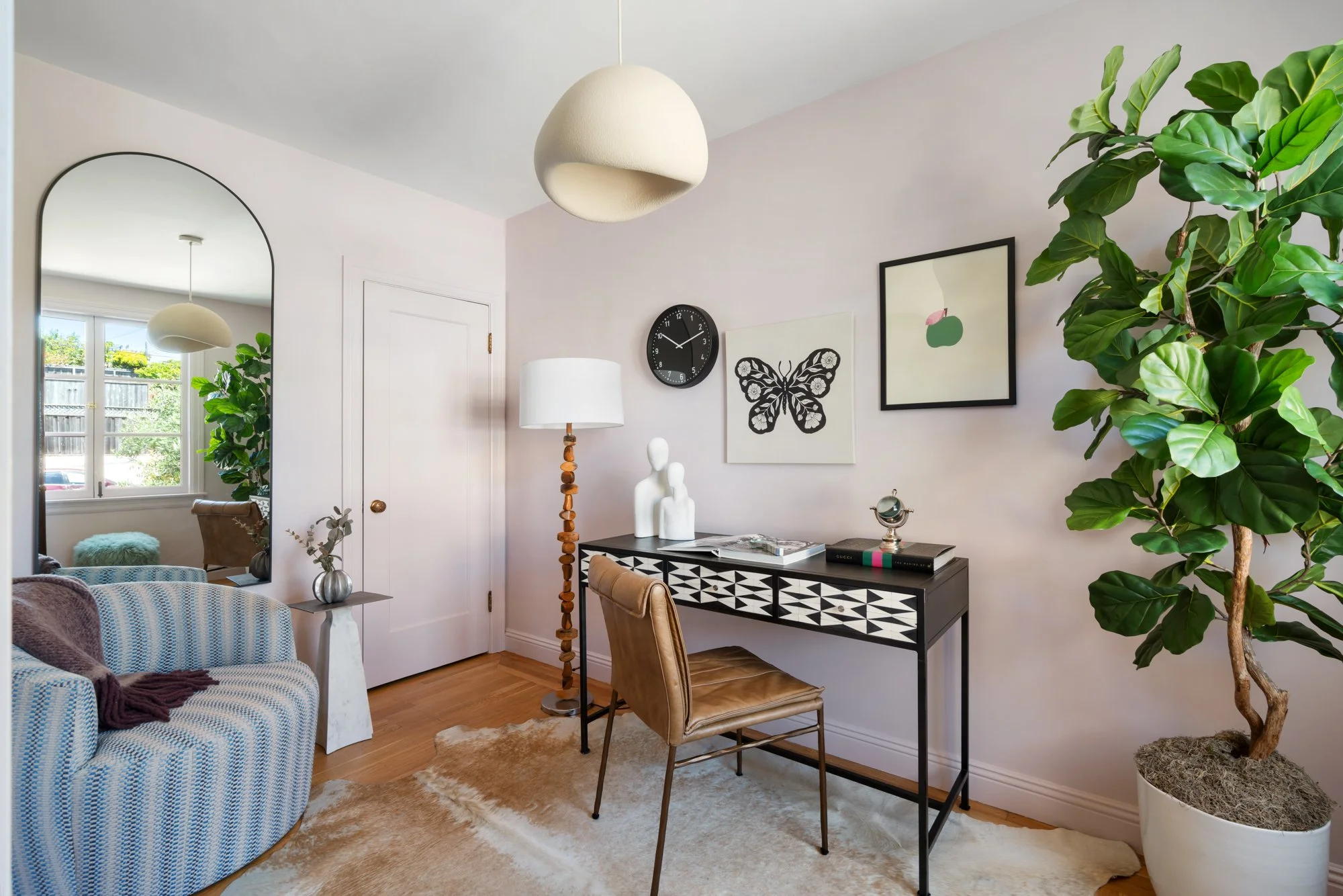 A cozy living room with a large potted fiddle leaf fig tree, a black and white patterned console table, wall art including a butterfly and apple print, a round black wall clock, a tall wooden floor lamp, a white door, and a mirror reflecting a window