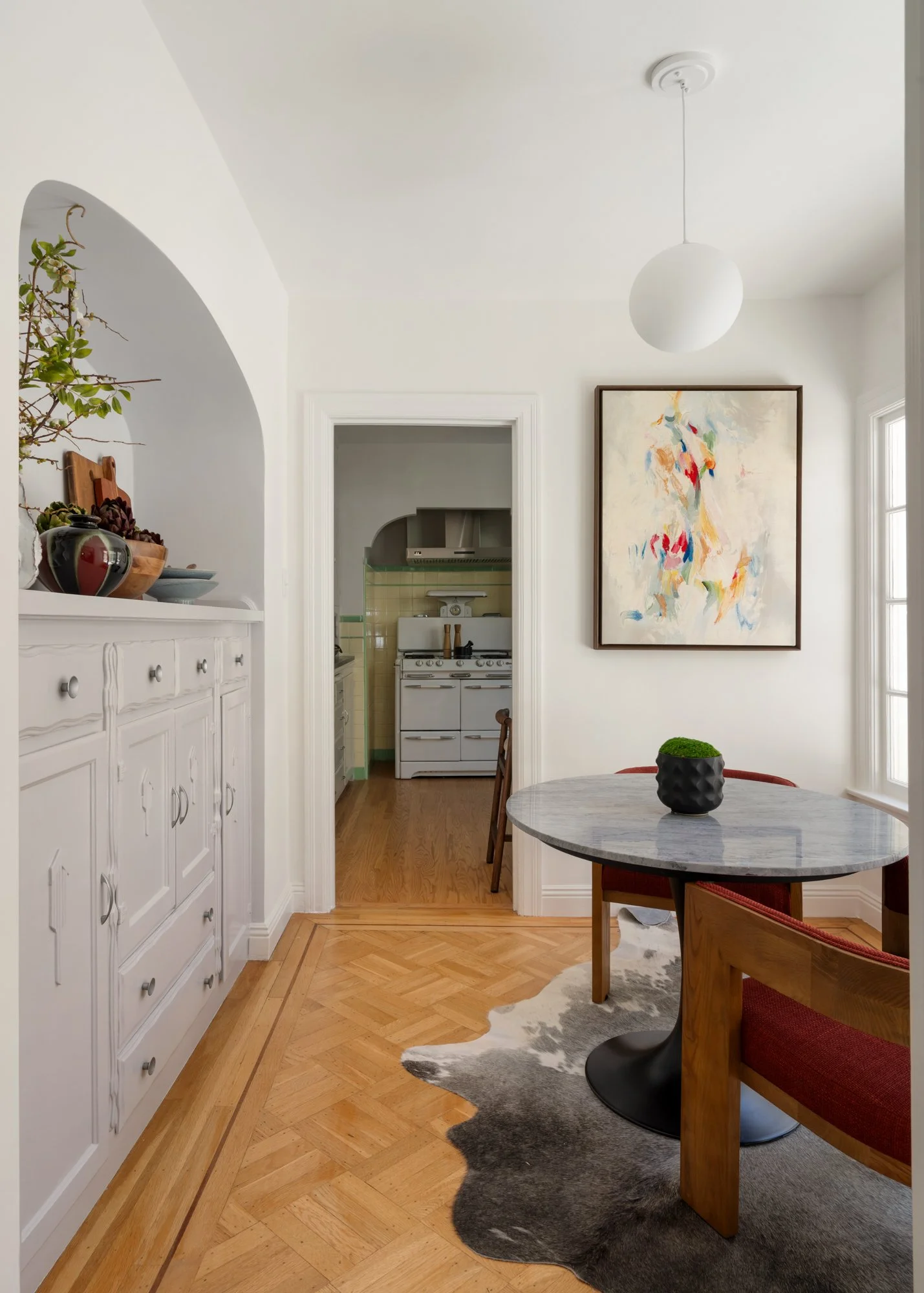 A cozy dining area with a round marble-top table, red upholstered chair, and black vase with green plant. White walls, abstract painting, window with sunlight, and wooden herringbone flooring.