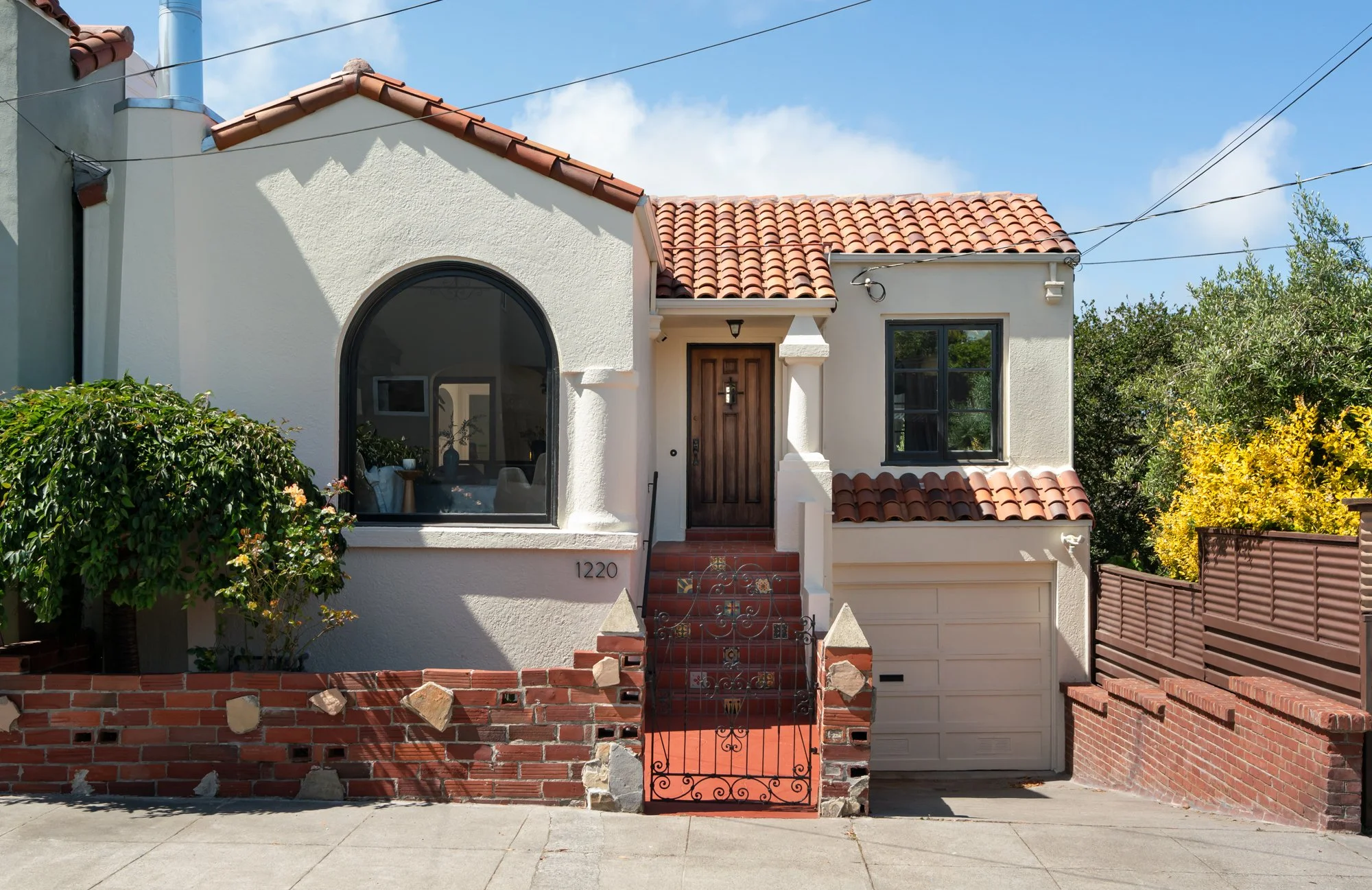 Front view of a white stucco house with a red tile roof, a large arched window, a wooden front door, a small staircase with iron railings, a brick wall with stones, greenery on both sides, and a driveway with a garage door.