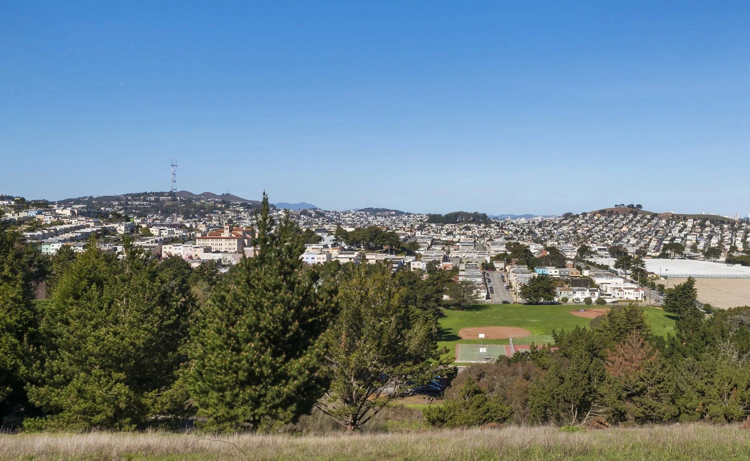 A panoramic view of a cityscape with residential buildings on hilly terrain, a communication tower on a distant hill, a park with trees and a baseball field, and a clear blue sky.