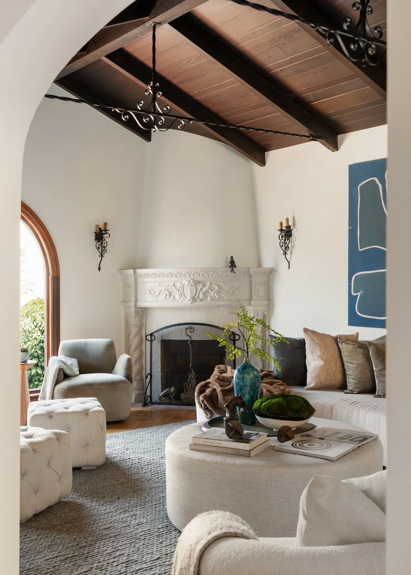 Living room with a beige tufted ottoman, a light-colored armchair, a plush sofa with beige and gray pillows, a fireplace with ornate trim, a blue vase with green branches, decorative objects, and a coffee table with magazines. Wooden ceiling beams an