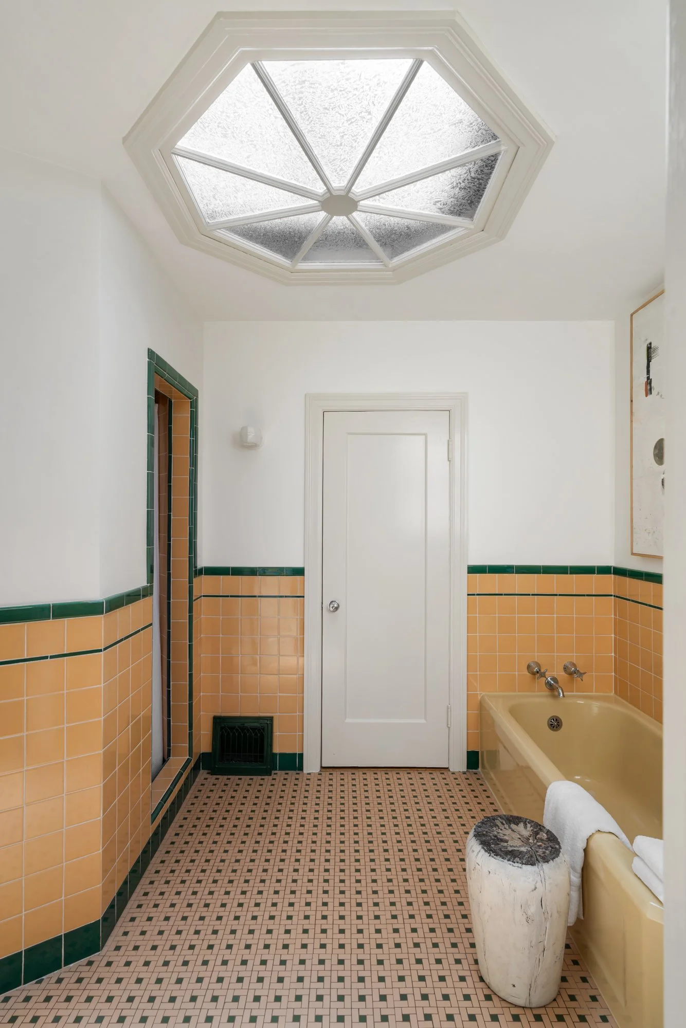 A bathroom with a decorative hexagonal skylight, yellow and green tiled walls, and a bathtub with a roll of toilet paper on a marble stand.