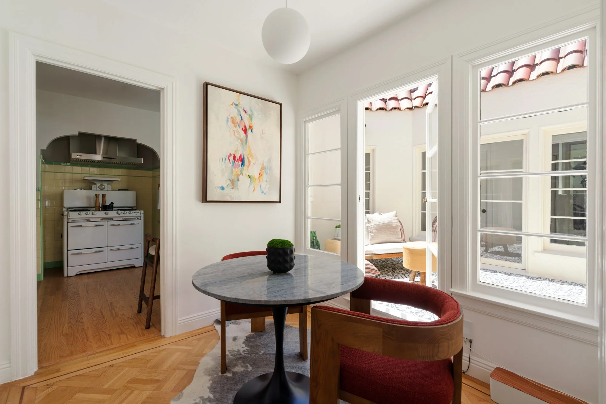 A cozy dining area with a round gray marble table and two wooden chairs with red cushions. A black vase with a small green plant sits on the table. Large windows behind with white frames and pinkish-red tiled roofs outside. An abstract artwork hangs 