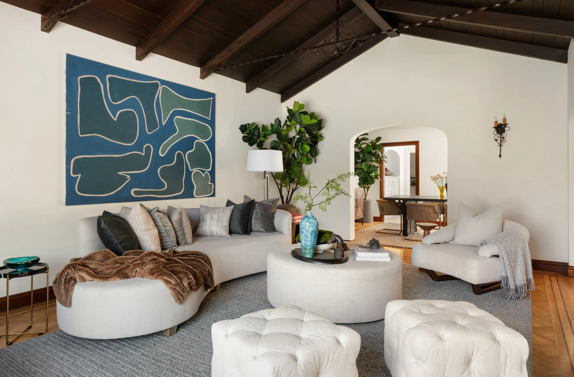 A living room with white sofas, accent pillows, a faux fur throw, a tufted ottoman, and a side table with a decorative bowl. There are large green houseplants, a blue abstract painting on the wall, and a wall-mounted candle holder with candles. The r