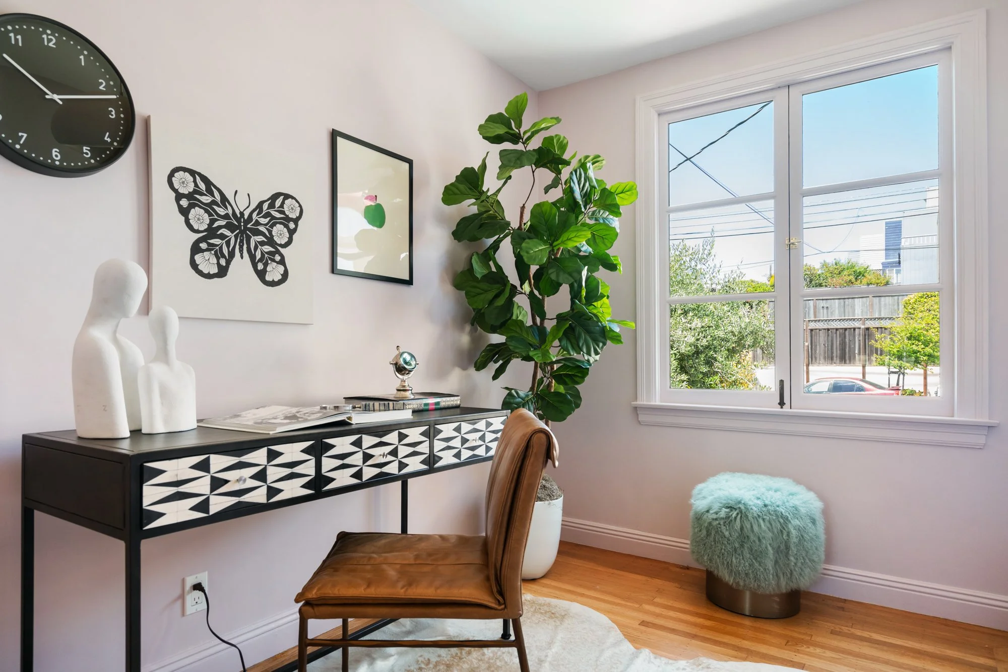 Interior view of a corner of a room with a black and white patterned side table, a brown chair with a leather seat, a large potted green plant near a window, decorative objects on the table including white sculptures and books, framed wall art featur