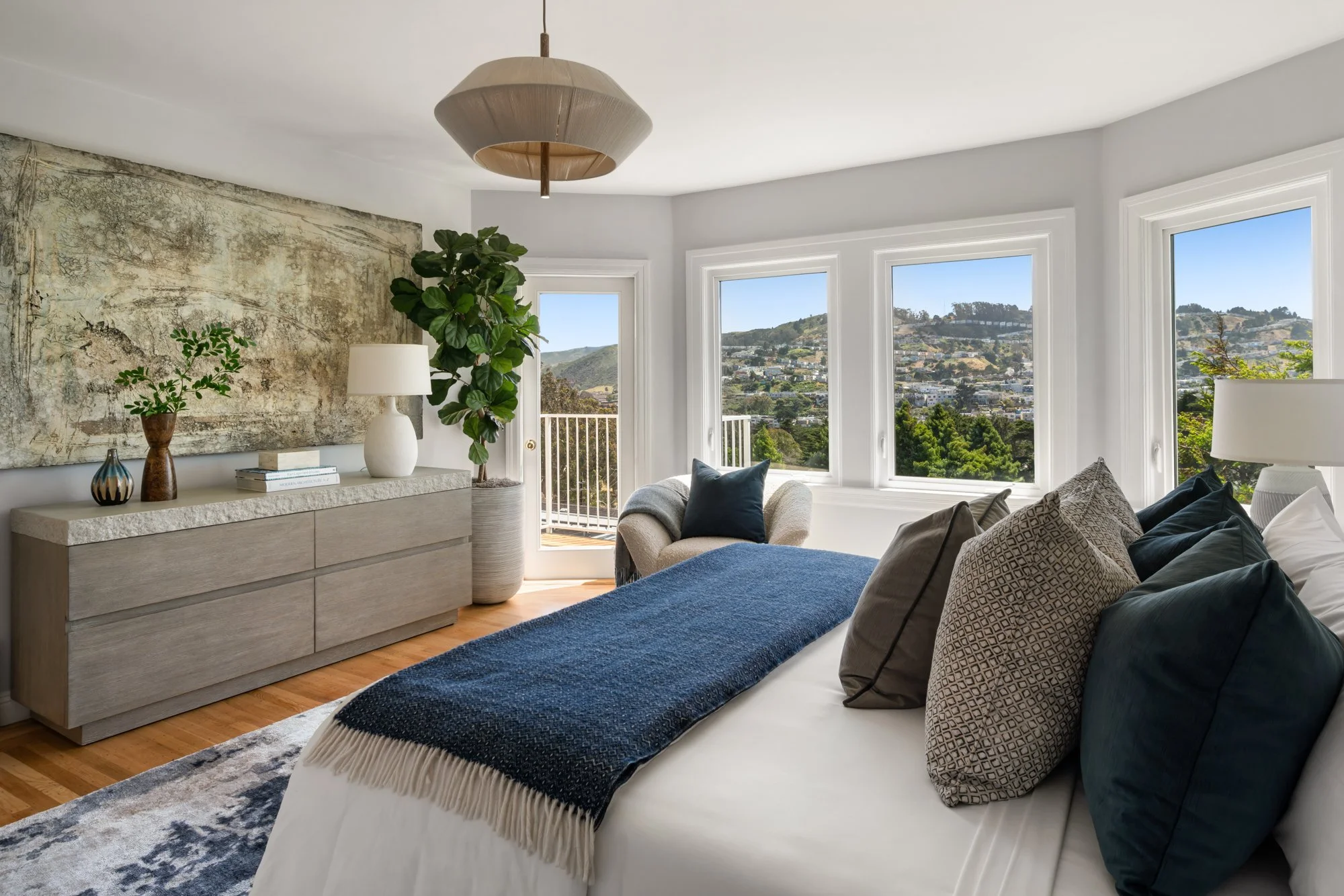 A bright living room with large windows showing a scenic view of hills and houses, a beige sideboard with decorative items, a tall potted plant, a beige armchair with pillows, and a blue throw on a sofa.