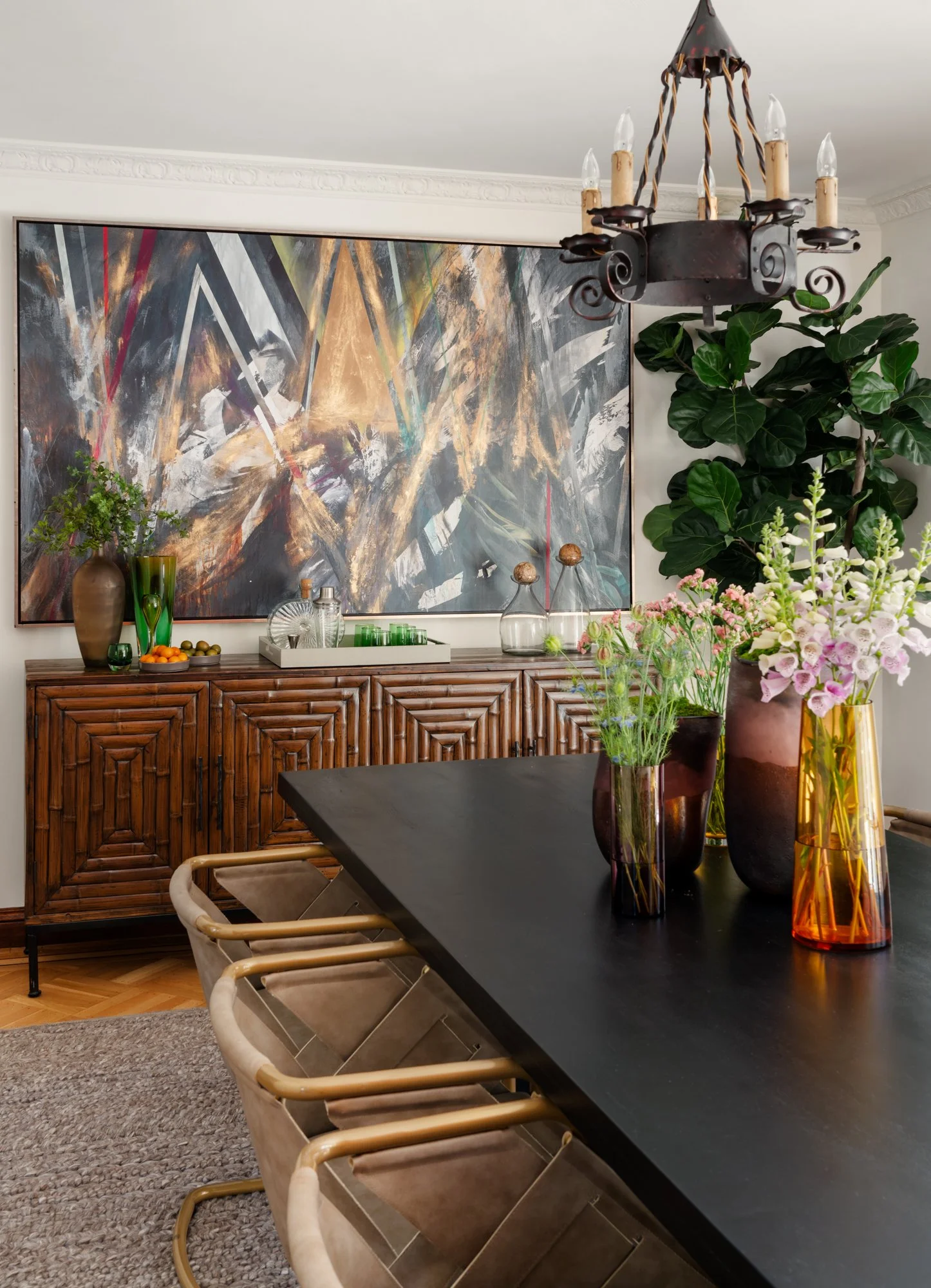A dining room with a black dining table decorated with orange and pink vases and various flowers. A large abstract painting hangs on the wall behind a wooden sideboard that holds glass and ceramic decor, including a large brown vase with greenery. A 