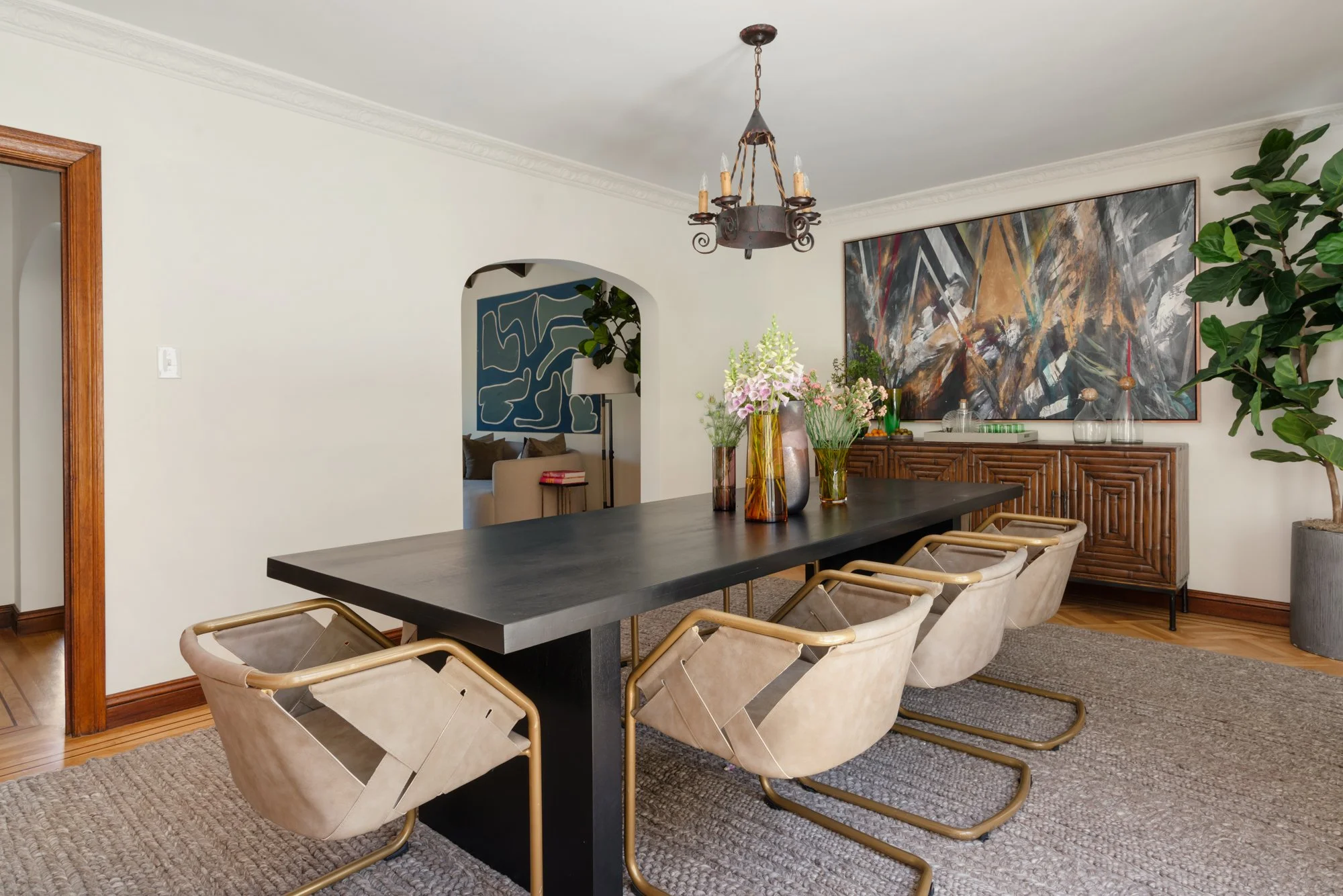 A dining room with a black table, beige chairs with gold legs, a large abstract painting on the wall, a chandelier, and a sideboard with decorative vases and plants.
