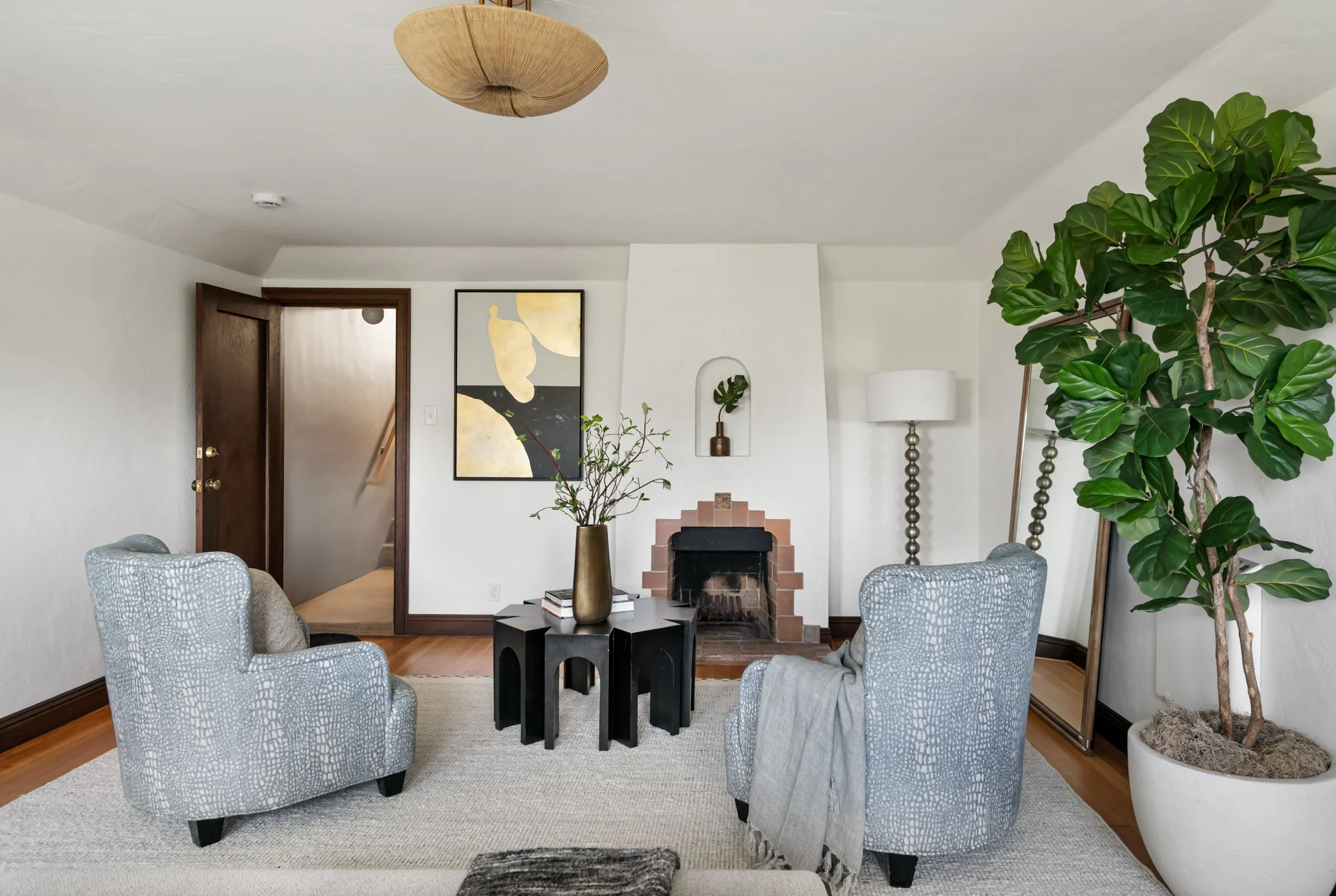 Living room with a fireplace, four patterned armchairs, a black coffee table with books and a vase, a large potted fiddle leaf fig tree, wall art, a floor lamp, and a doorway leading to a staircase.