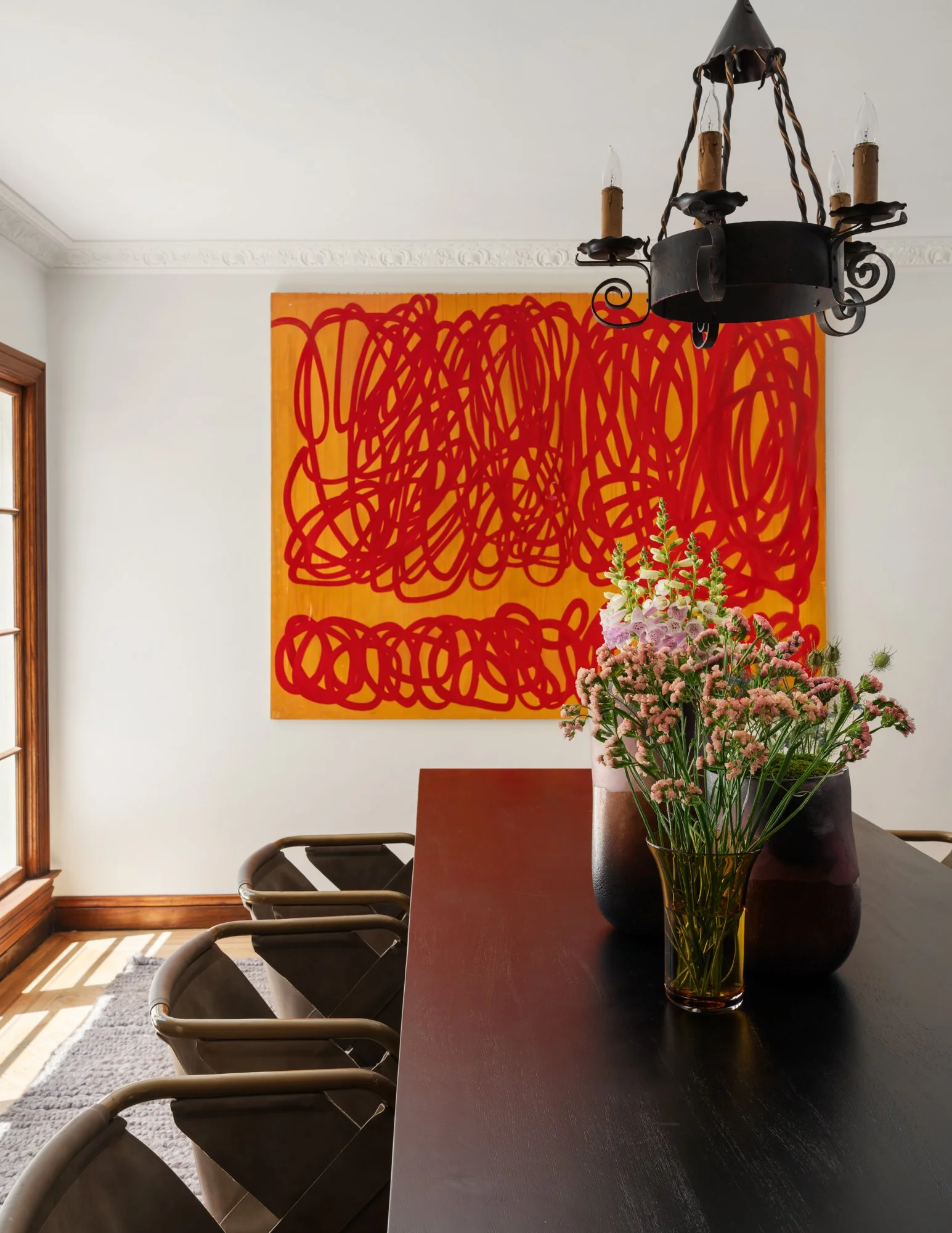 A dining room with a dark wood table, four modern chairs, a black vase with pink flowers, a yellow vase, a smaller dark vase, a chandelier with four candle-shaped bulbs, and a large abstract red and orange painting on the white wall.