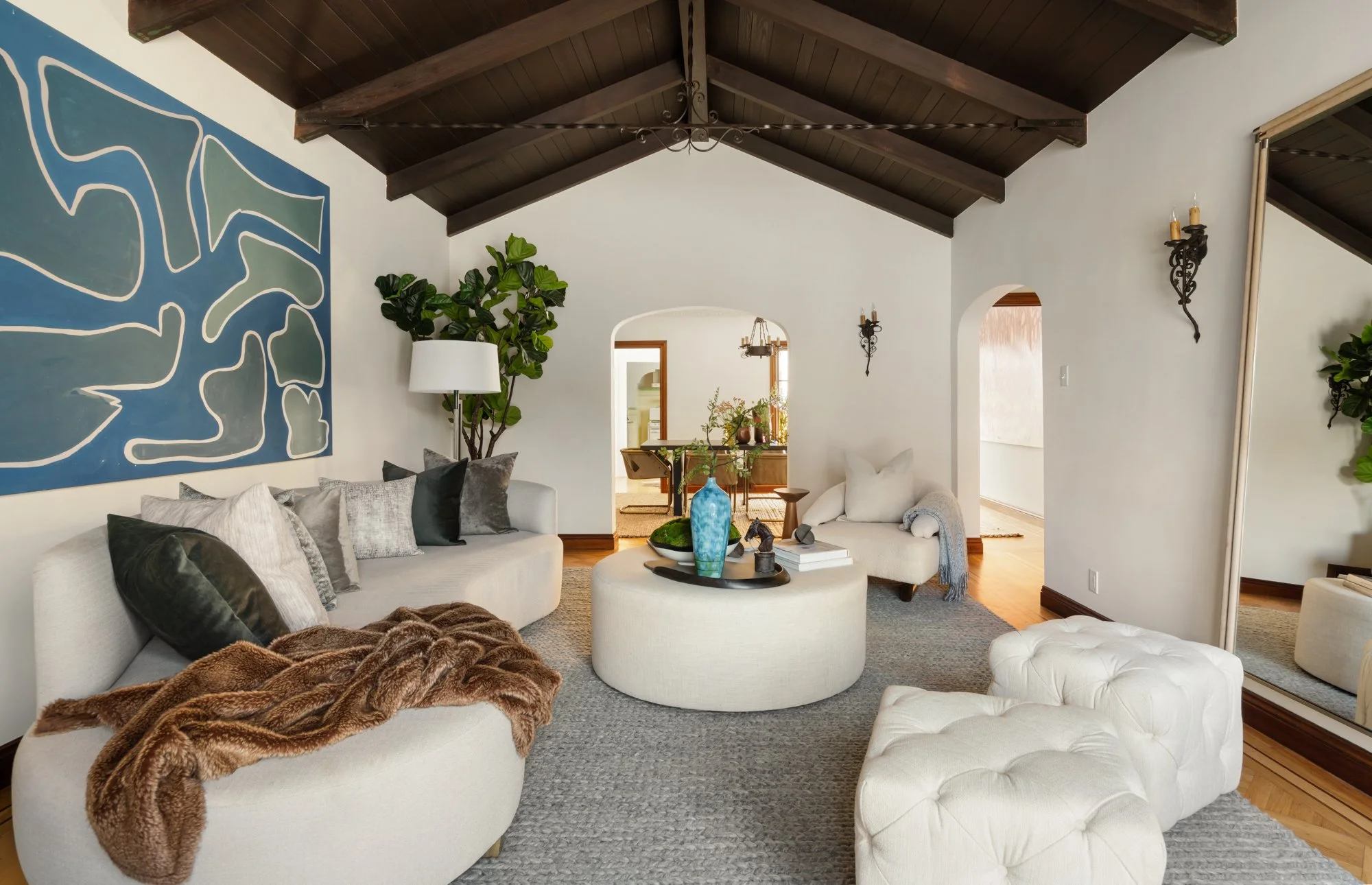 Living room with white sofas, decorative pillows, a fur throw, a round ottoman, a large abstract wall art, a potted plant, a blue vase, and wall-mounted lanterns, with a view into a dining area and kitchen.