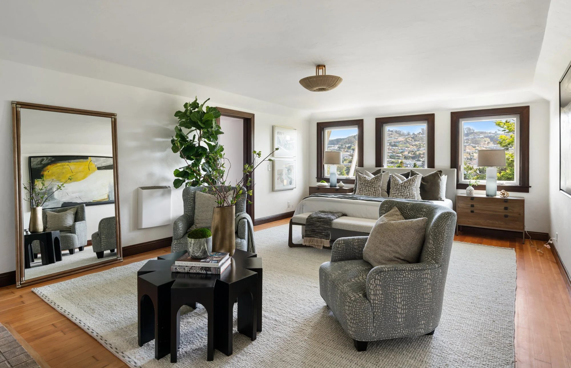 Living room with large window view, neutral-colored furniture including armchairs and a bed, decorative items on side tables and a mirror, and indoor plants.