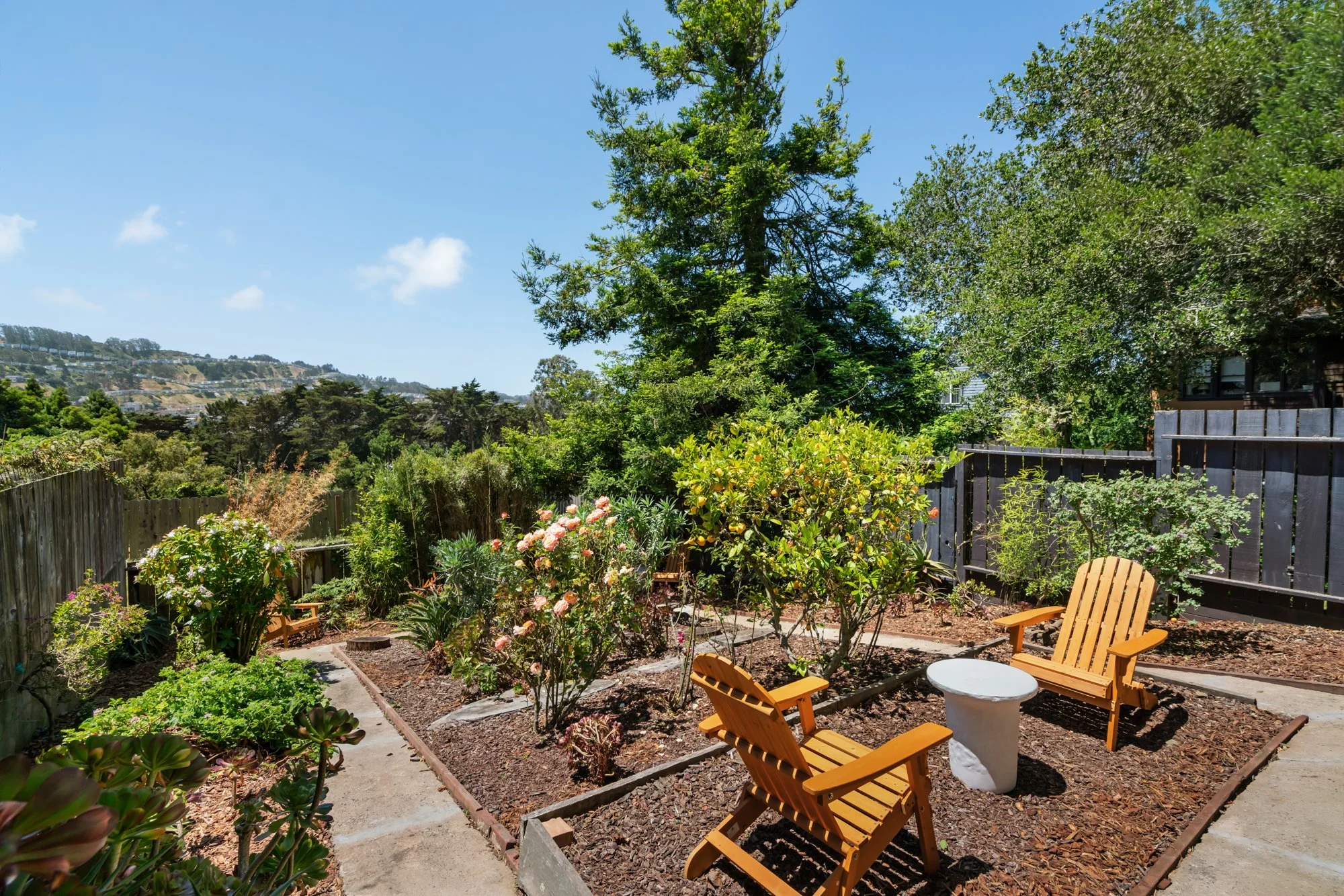 A backyard garden with wooden chairs and a small round white table on the patio, surrounded by various flowering plants, trees, and shrubs, with a fence and hills in the background on a sunny day.