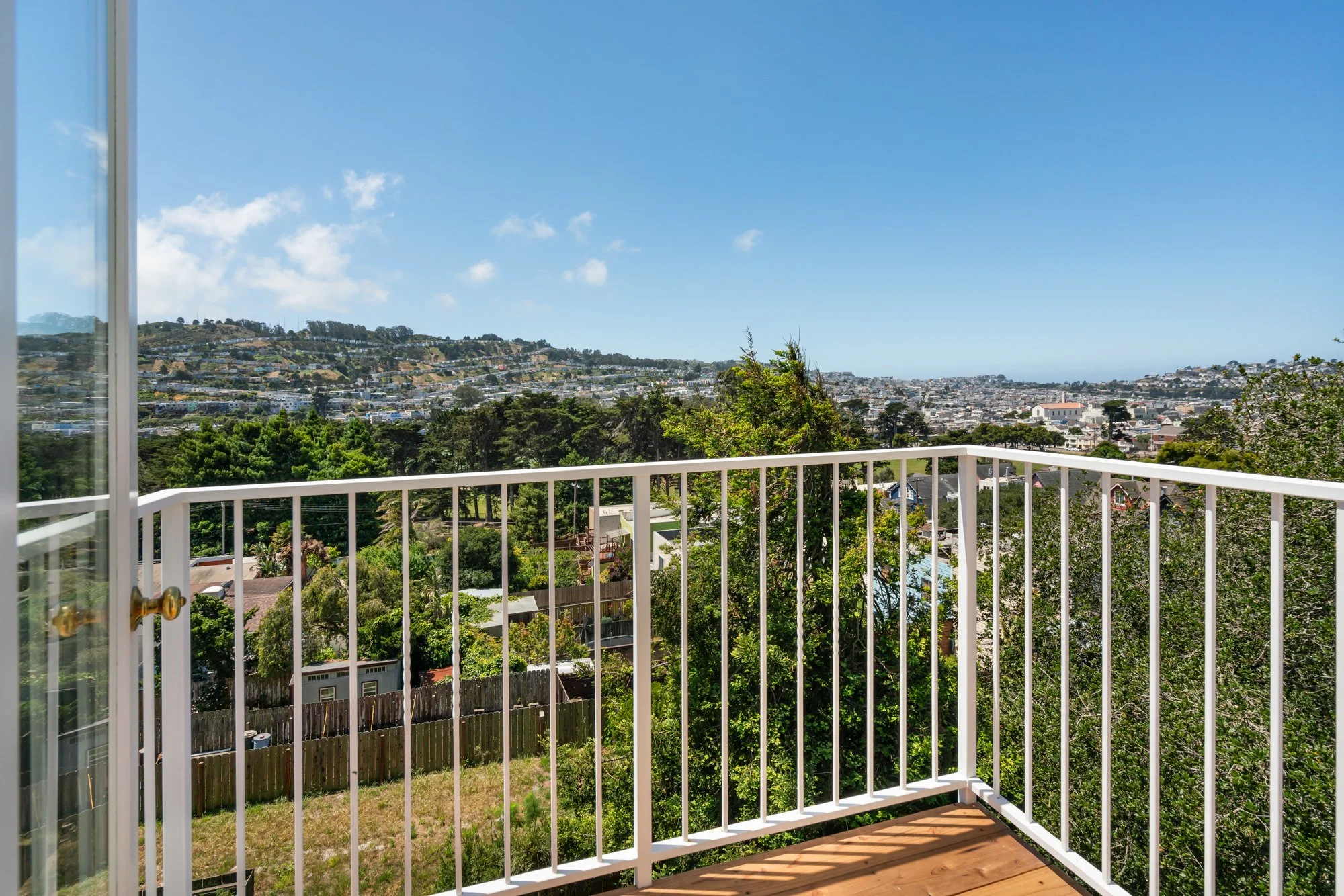 View from a balcony showing a suburban area with hills, houses, and lush green trees under a clear blue sky with a few clouds.