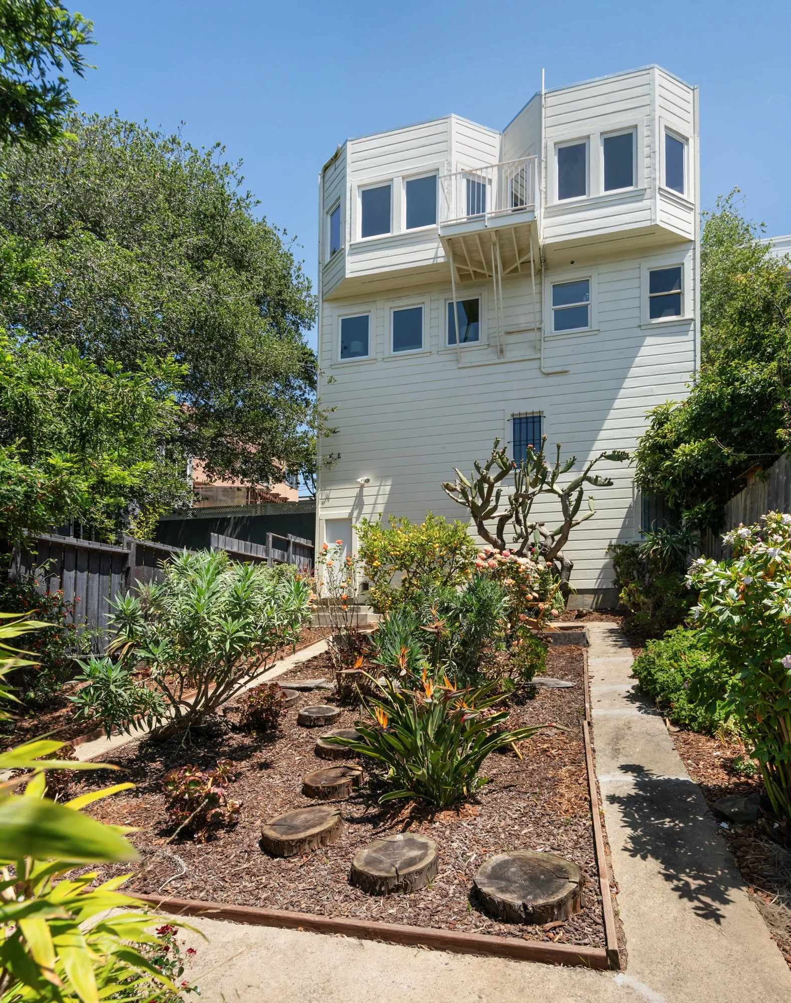A white multi-story house with a garden and a stone pathway. The garden has various plants and bushes, and there is a wooden fence. The house features multiple windows and a small balcony.