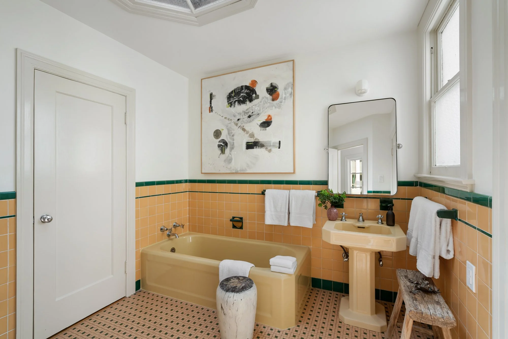 A vintage bathroom with yellow bathtub, yellow sink, and peach-colored tile walls with green accents. The bathroom features a large abstract artwork on the wall, a large mirror, windows, and various towels and decorative items.