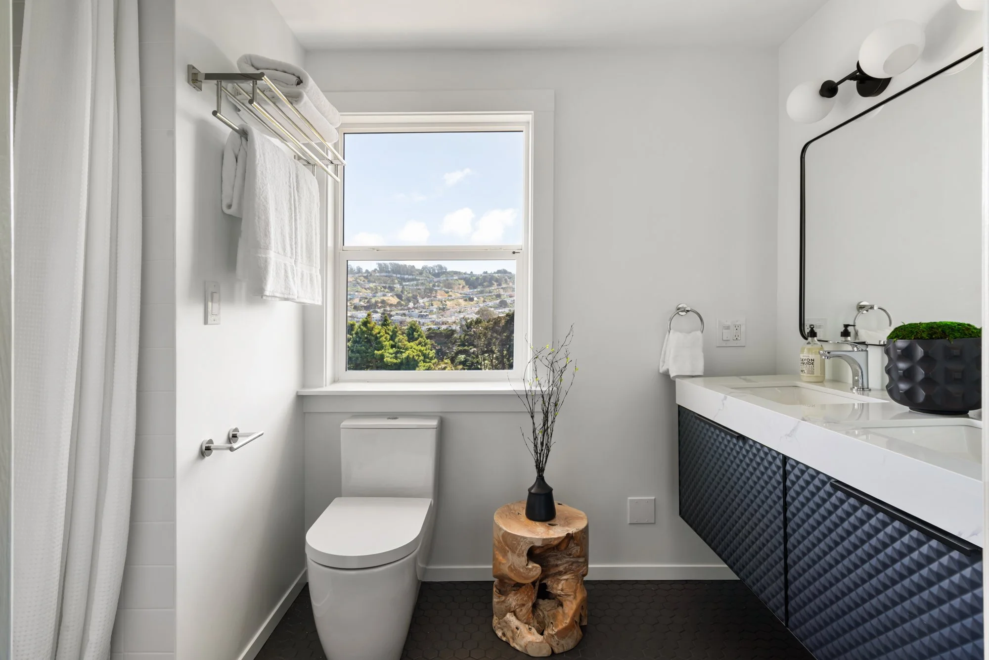 Minimalist bathroom with white walls, large window showing a city view, black hexagonal floor tiles, and black vanity with geometric textured cabinet doors. Decor includes a black vase with branches, wall-mounted towel rack with white towels, and bla