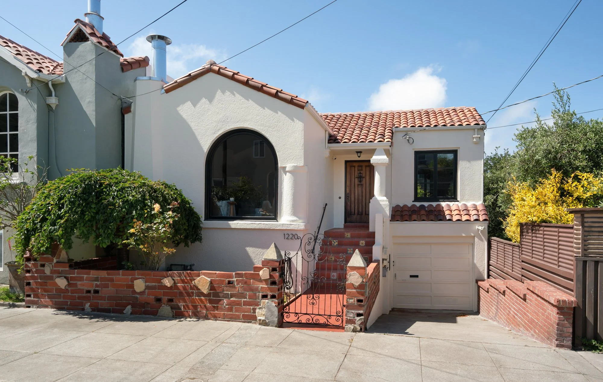 Front view of a white stucco house with a red tile roof, a large arched window, and a wooden front door. There is a small brick, wrought iron, and concrete fence with a gate at the front. Some green bushes and yellow flowering plants are in the yard.
