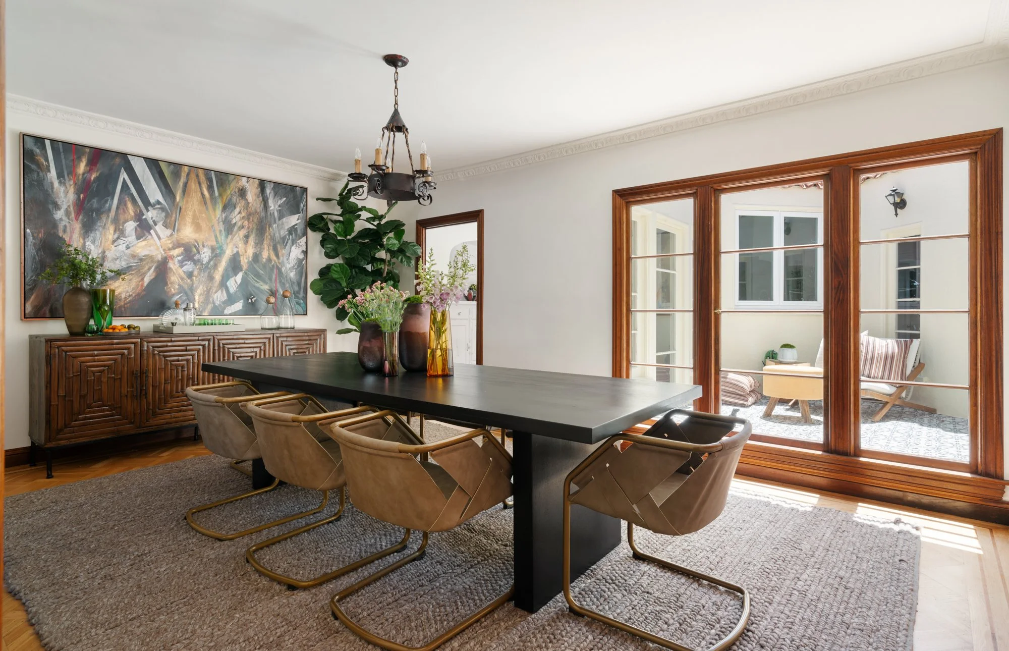 A dining room featuring a large black rectangular table surrounded by beige cushioned chairs with gold frames. The room has a textured beige carpet and a wooden sideboard decorated with vases and a bowl of fruit. On the wall, there is a large abstrac