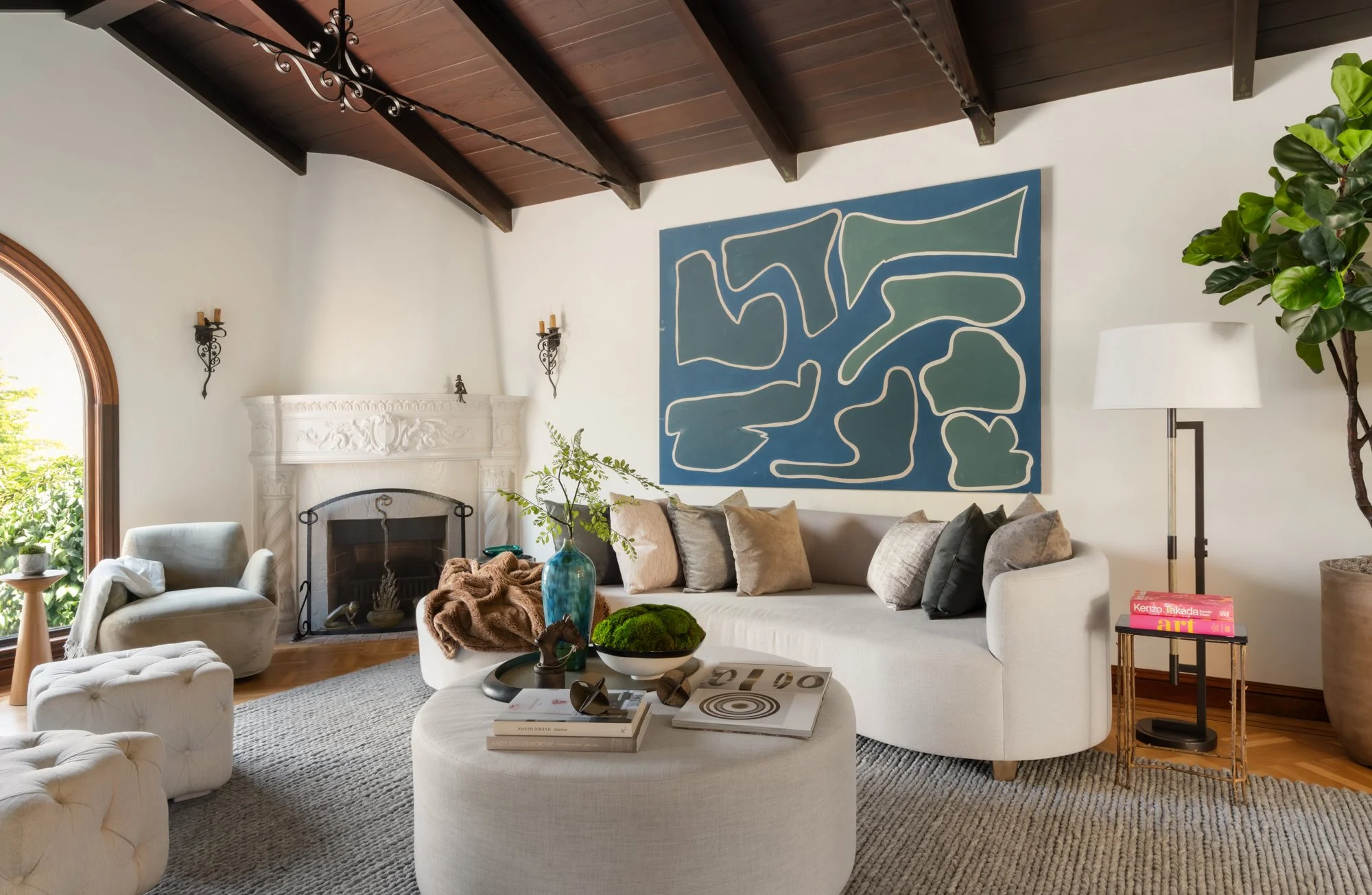 Living room with white walls, a wood-finished ceiling, a fireplace, a large abstract blue-green wall art piece, a beige sofa with pillows, a white tufted ottoman with books and decorative items, a comfortable armchair with a blanket, a large green pl