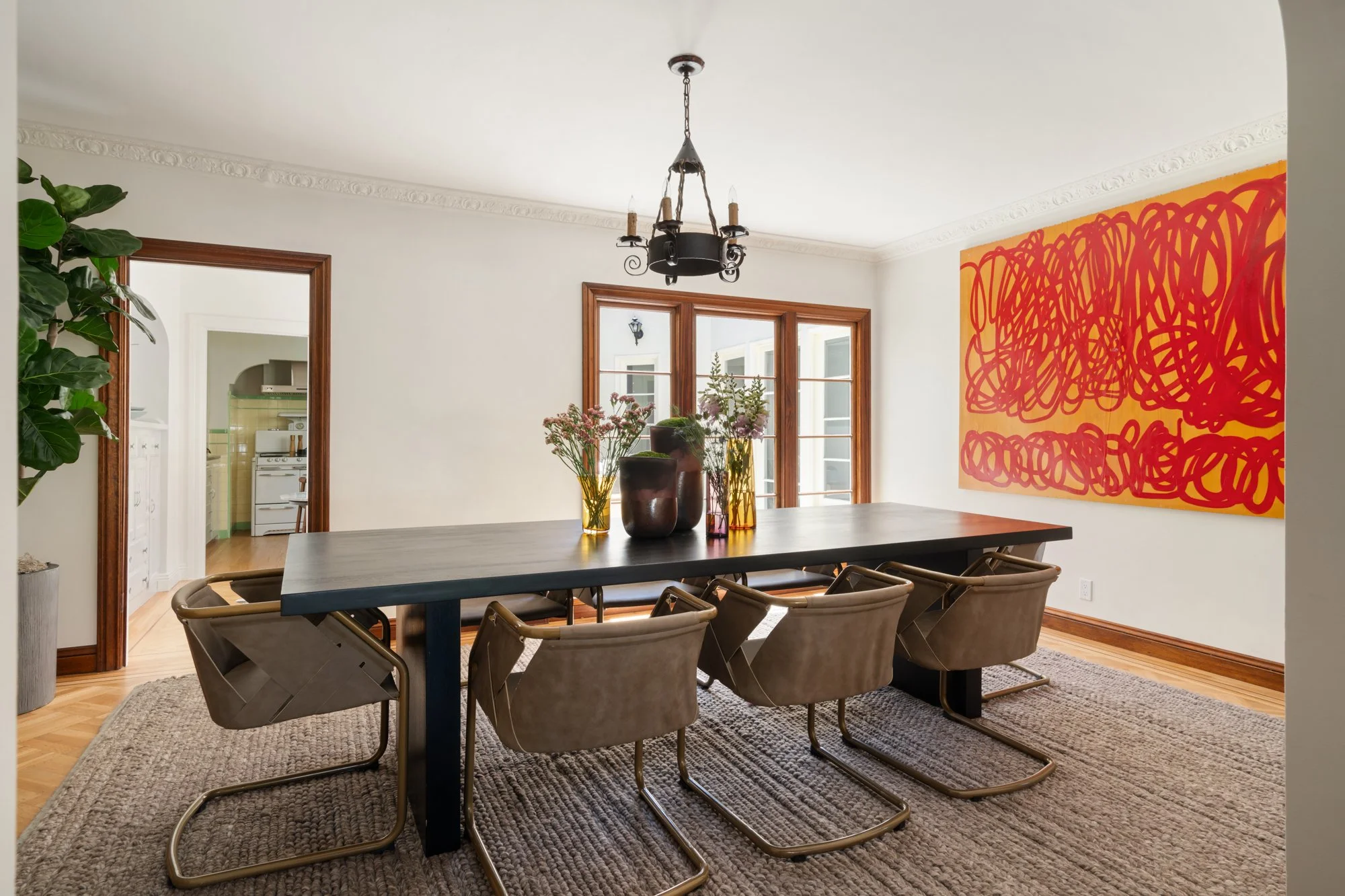 A dining room with a long black table, beige chairs, and vases with flowers. A large abstract red and yellow painting hangs on the wall, with windows and a chandelier overhead.