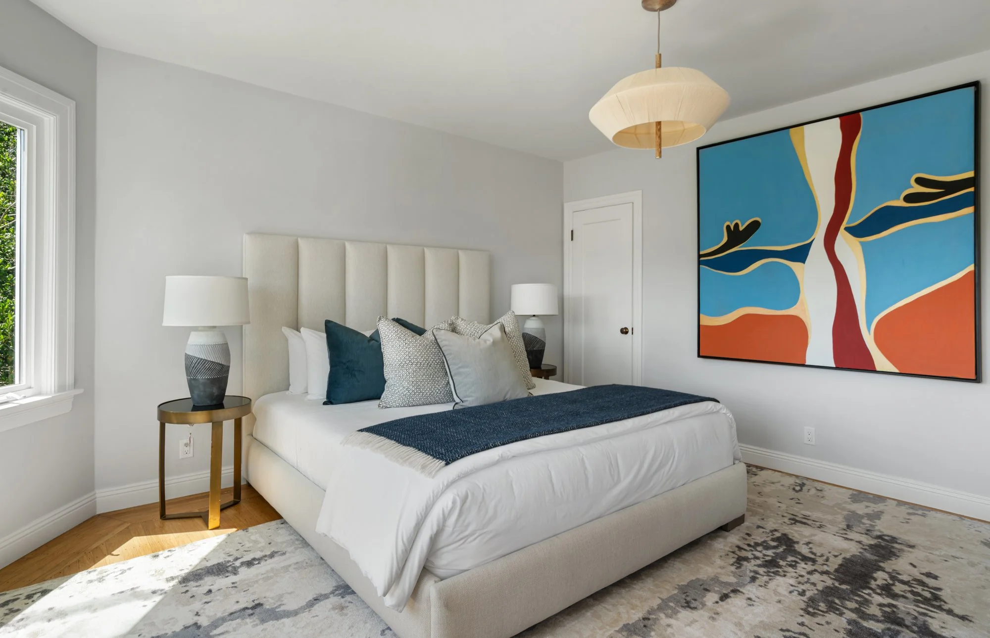 Modern bedroom with white upholstered bed, decorative pillows, a navy blue throw blanket, one side table on the left with a patterned ceramic lamp, an abstract colorful painting on the wall, and a window showing greenery outside.