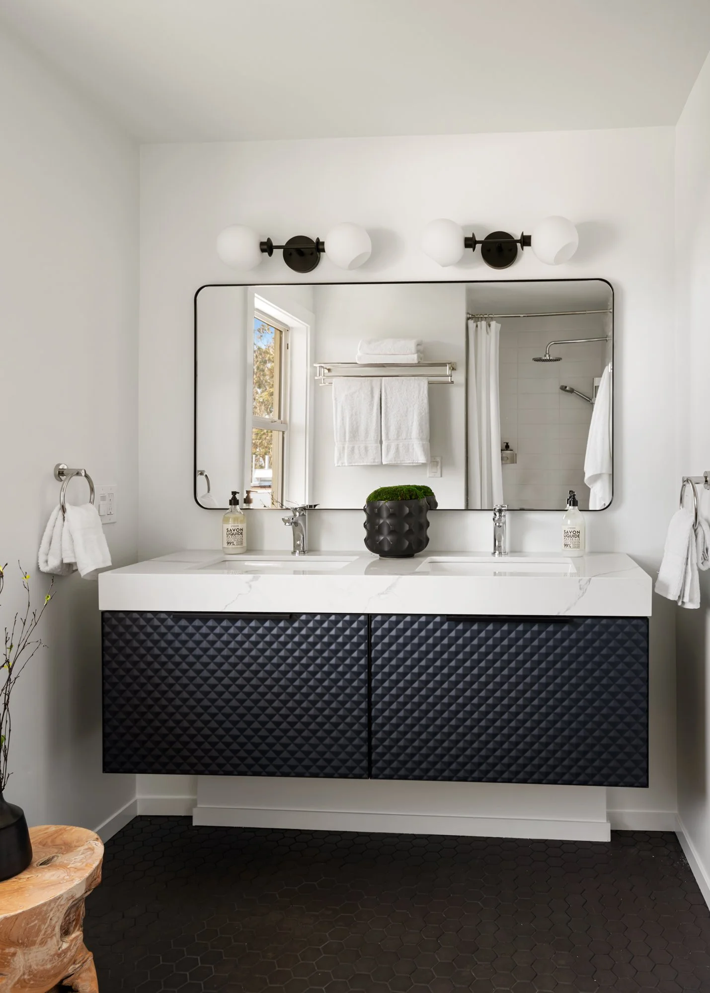 A modern bathroom with a black vanity featuring textured cabinets, a white marble countertop with two sinks, a large mirror, and two wall-mounted light fixtures with white globe shades. Towels hang on towel rings on either side, and a small window re