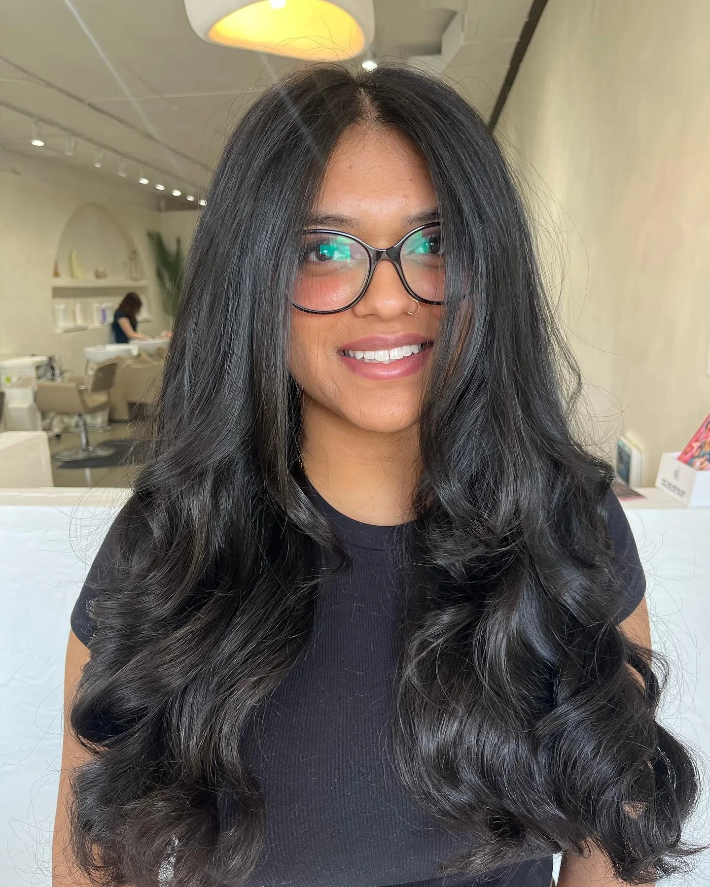 A woman with long, wavy black hair, wearing glasses and a black shirt, smiling in a well-lit salon or spa setting.