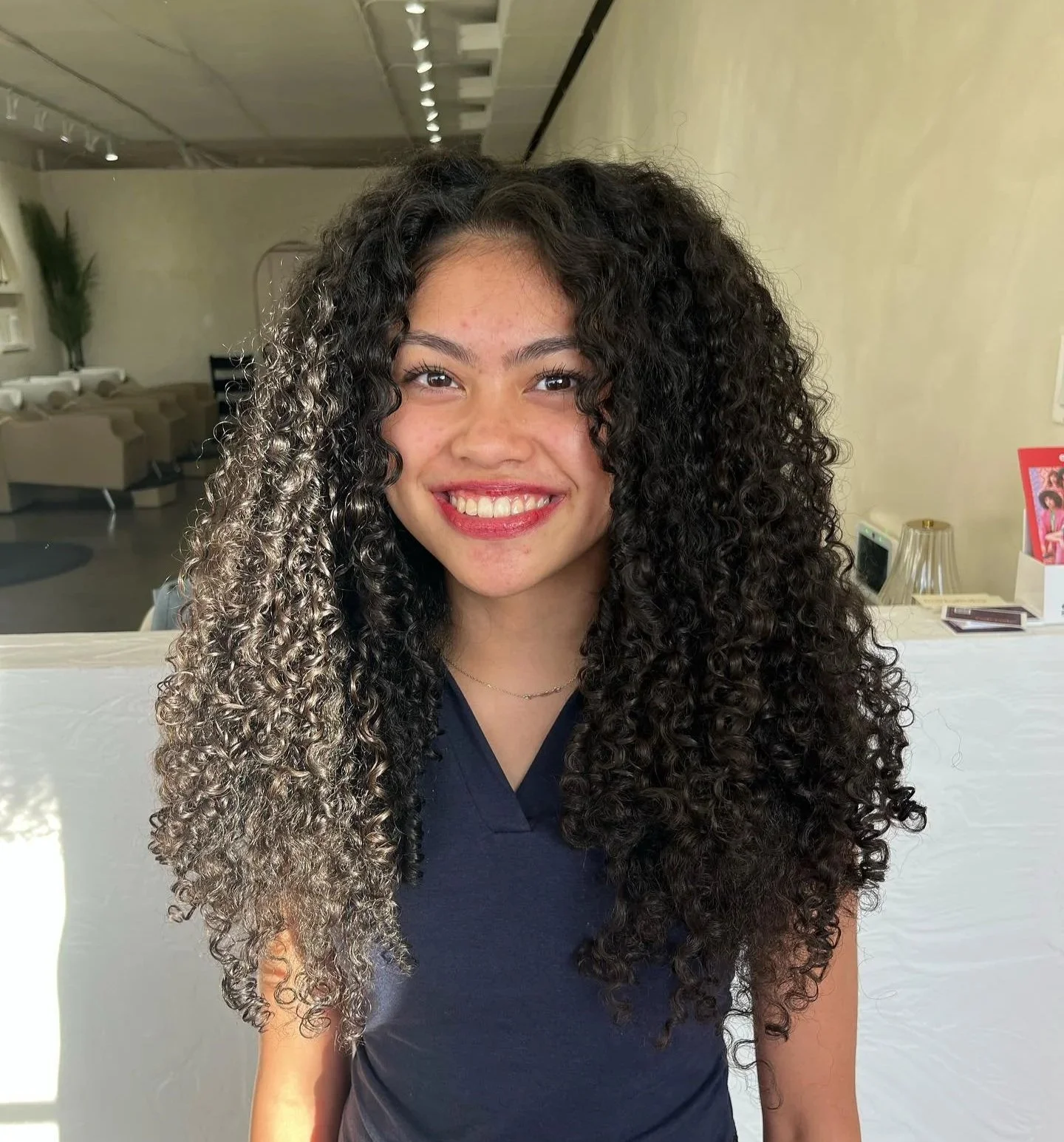 Young woman with curly hair, smiling, wearing a Navy blue top, sitting indoors with a modern decor background.