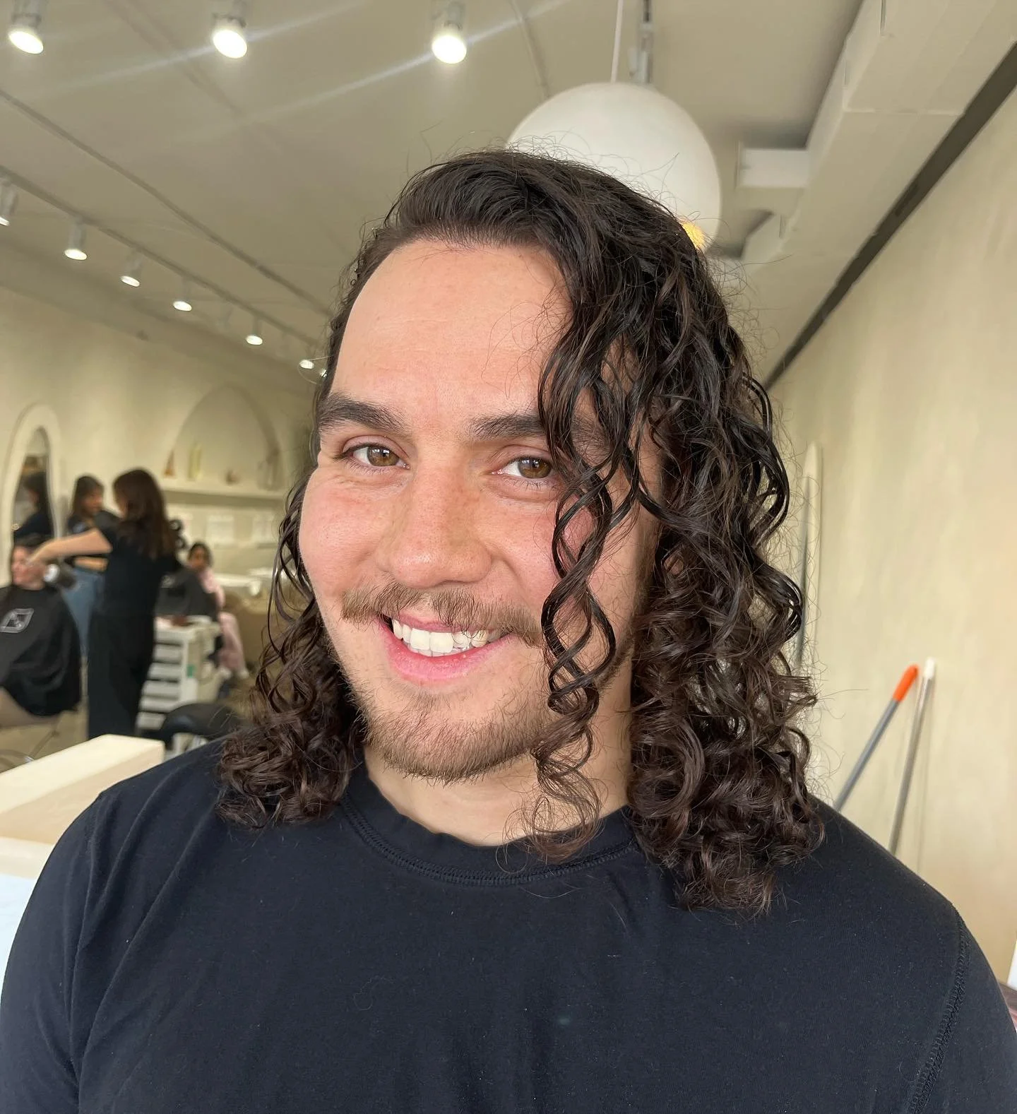 Smiling man with long curly hair, wearing a black shirt, in a salon or studio with people and hair styling chairs in the background.