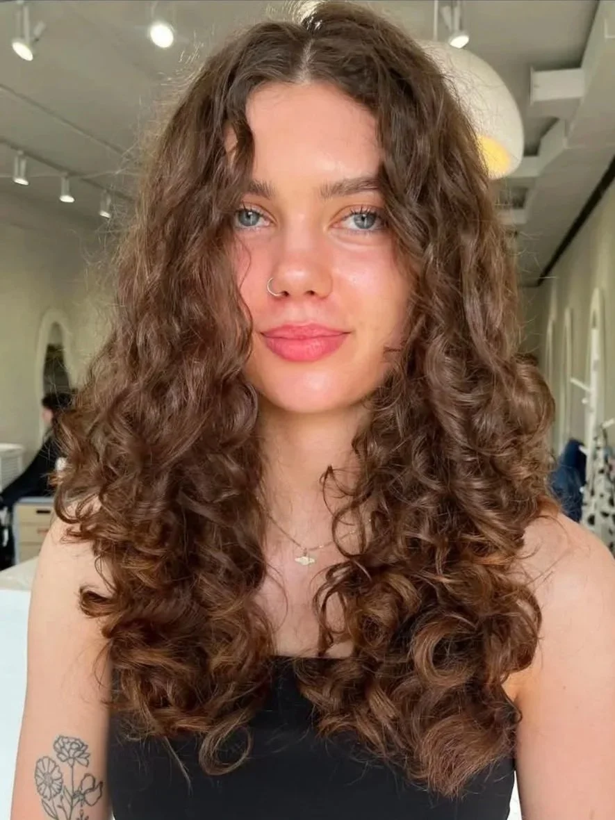 A young woman with curly brown hair, light skin, and blue eyes, wearing a small nose ring, a necklace with a cloud pendant, and a black top, posing indoors with a neutral expression.