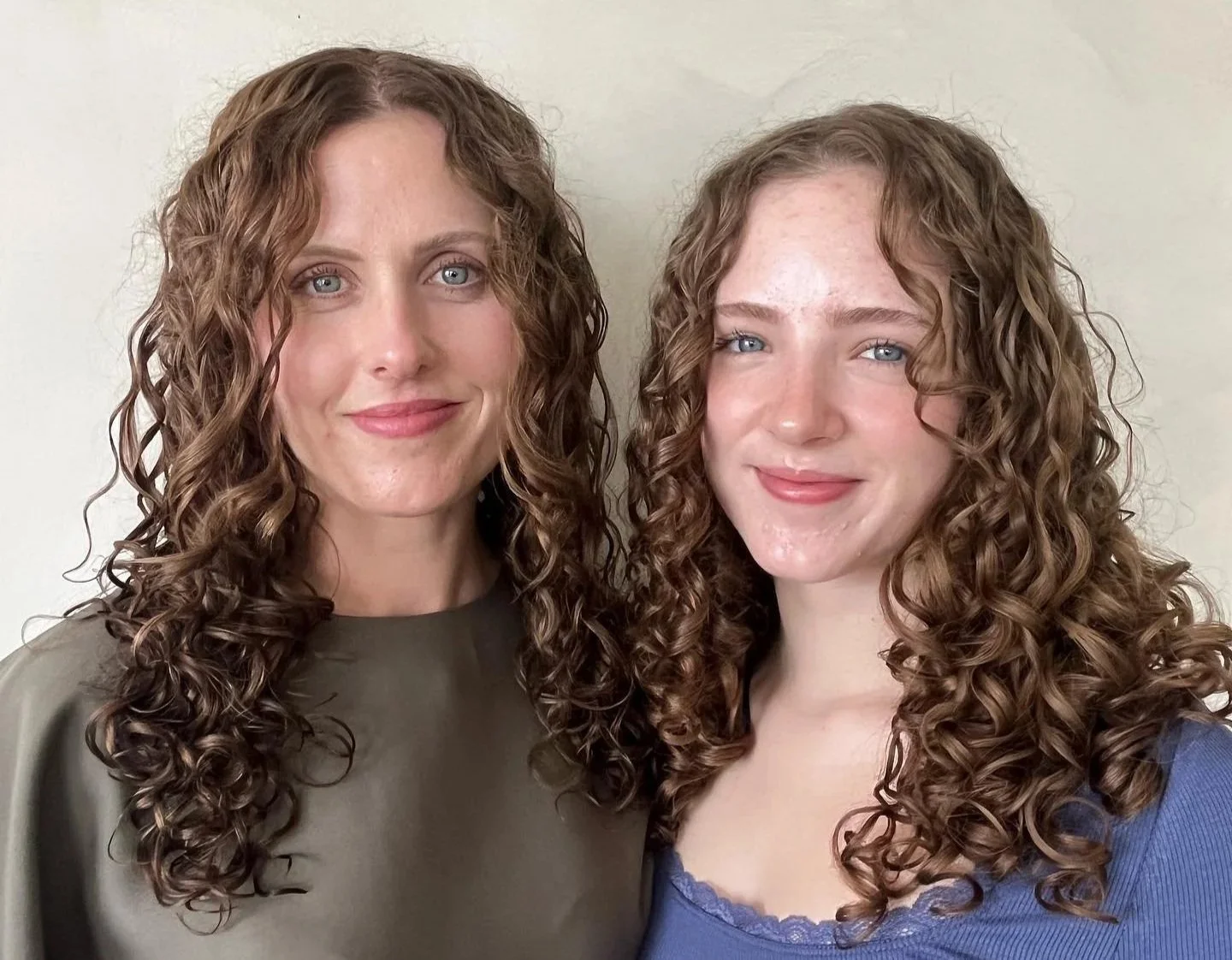 Two women with curly brown hair and blue eyes smiling at the camera, standing against a plain light-colored wall.