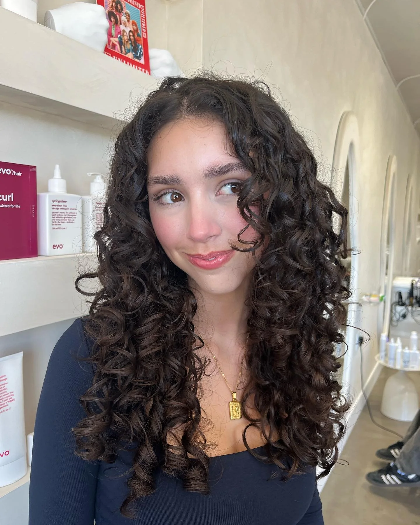 Young woman with long, curly dark hair, wearing a black top and gold necklace, smiling in a salon or stylist's space.