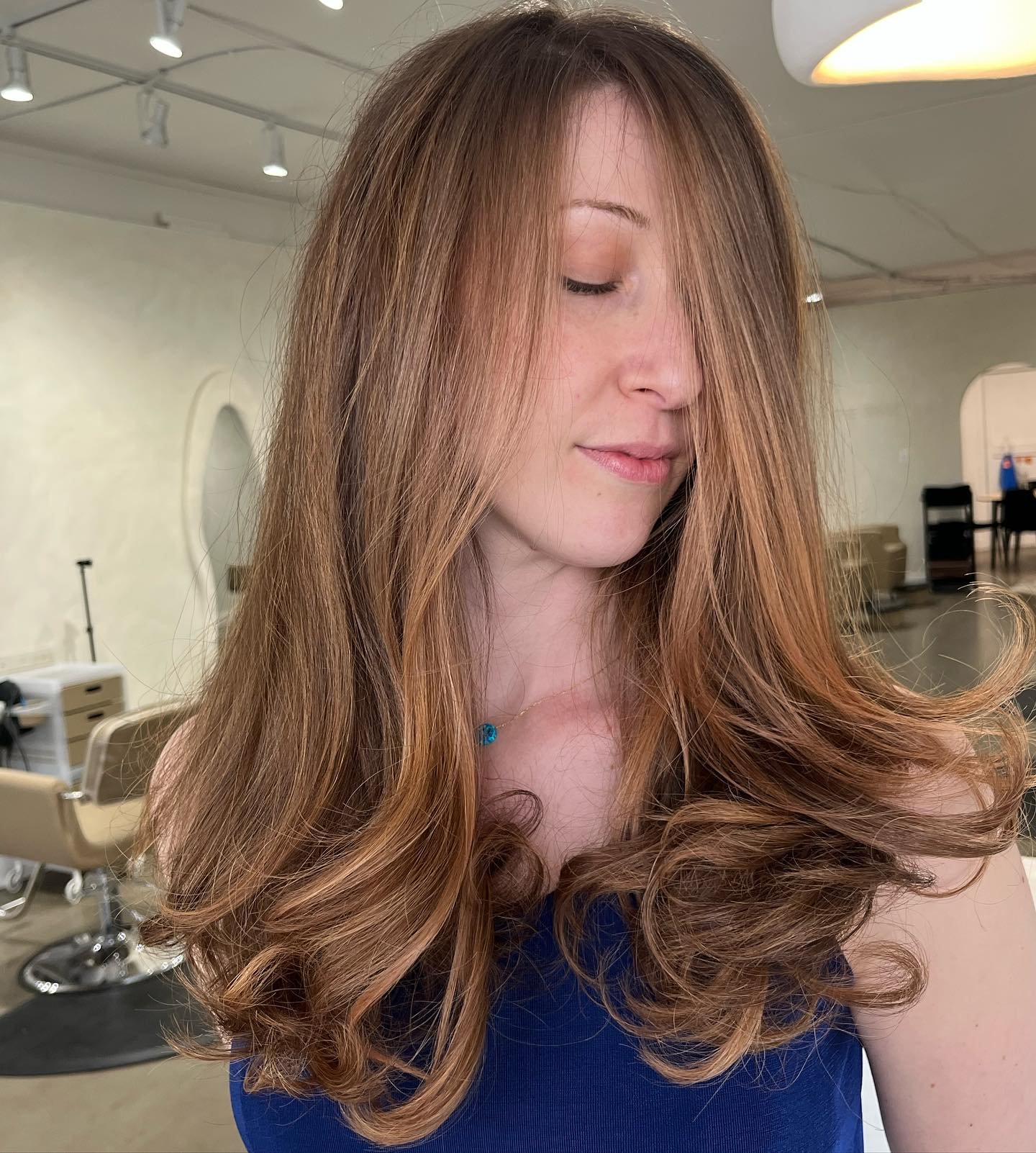 A woman with long, wavy, reddish-brown hair and fair skin, with her eyes closed and a gentle facial expression, inside a hair salon.