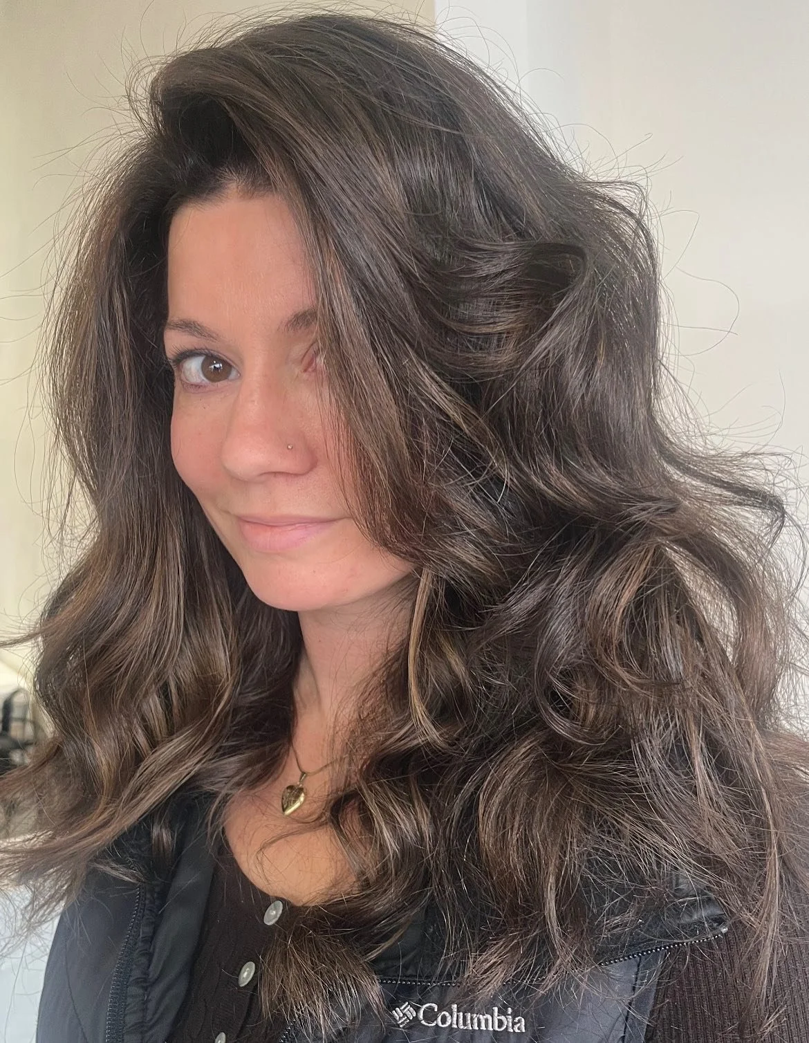 A woman with long, wavy brown hair taking a selfie indoors. She is wearing a black Columbia jacket and a gold heart necklace.