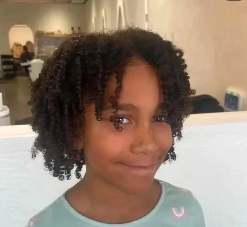 A young girl with natural curly hair smiling in an indoor setting.