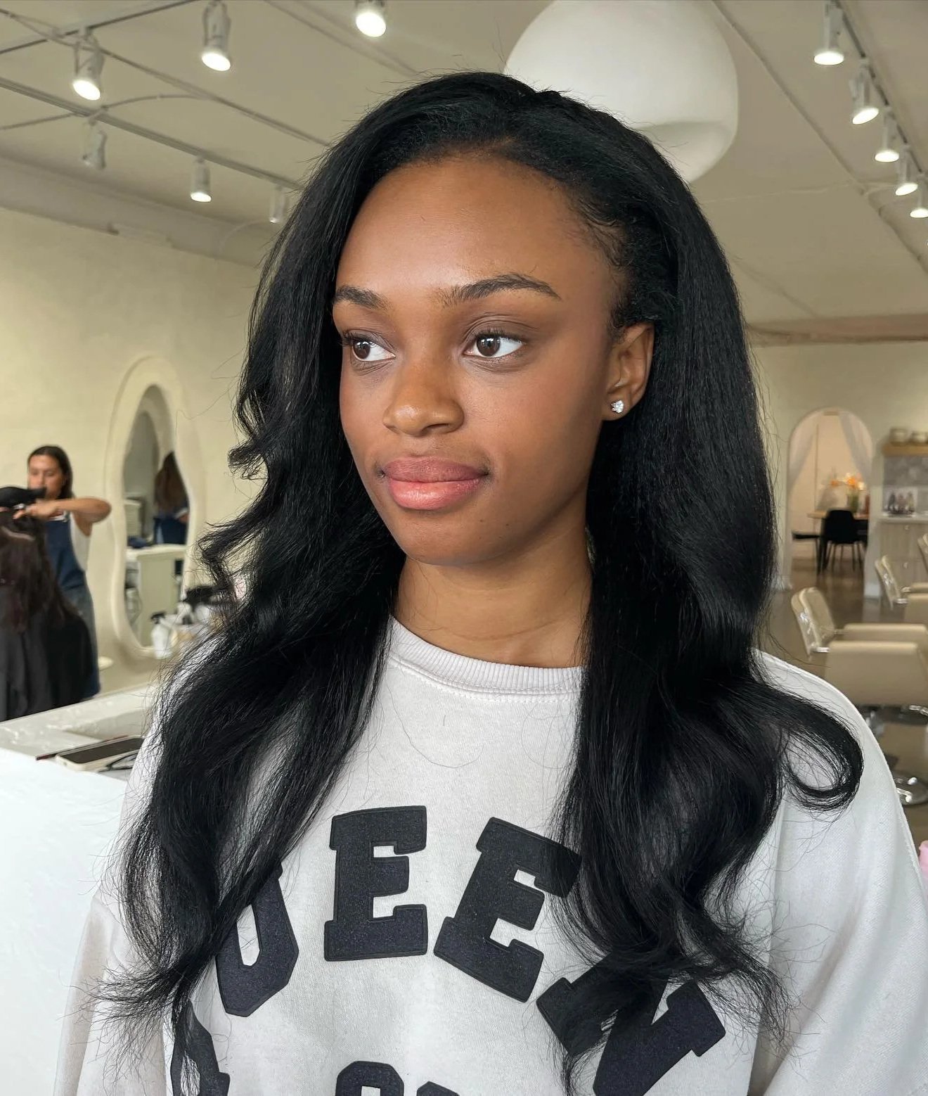 Young woman with long black hair and diamond stud earrings in a salon, wearing a white sweatshirt with black letters