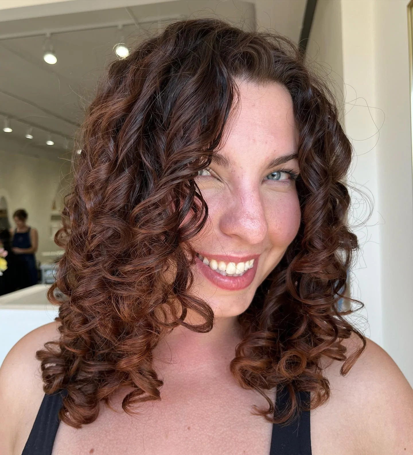 Close-up of a woman with curly brown hair smiling and winking, wearing a black tank top. Indoor setting with bright lighting.