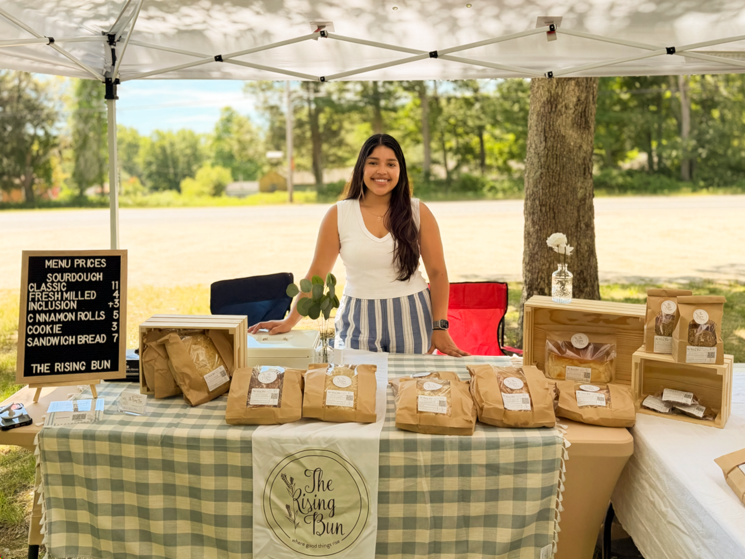 A woman stands behind a table displaying bags of baked goods and a small sign with menu prices at an outdoor market stall. She is smiling and wearing a white top and striped apron. A large white canopy tent is overhead, and trees recess in the background.