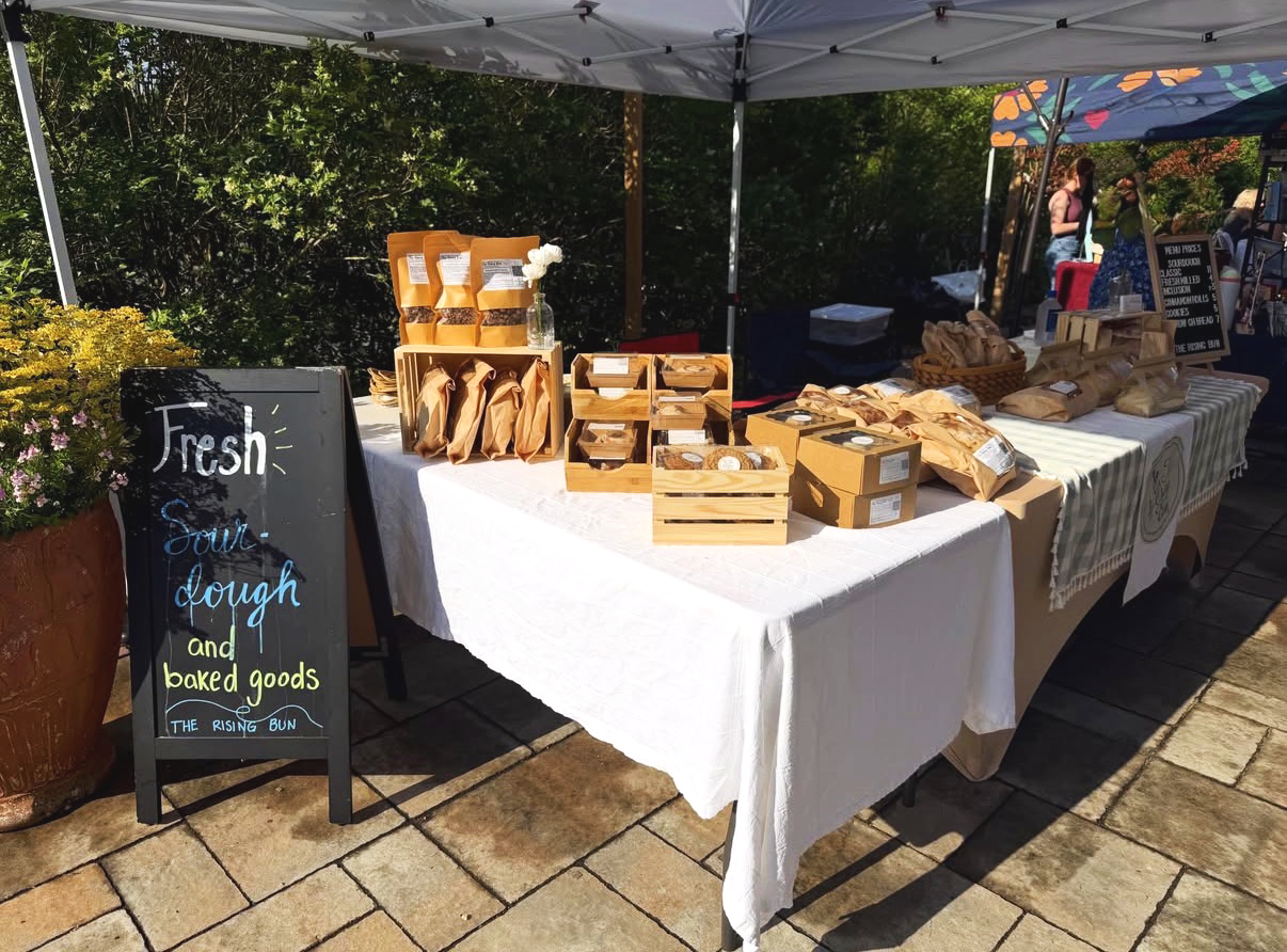 A farmers market stall selling sourdough and baked goods. The stall has a white tablecloth and is shaded by umbrellas. There is a black chalkboard sign that reads "Fresh sourdough and baked goods" with a smaller sign underneath. Baked goods are displayed in wooden boxes and packages. In the background, there are people and trees.