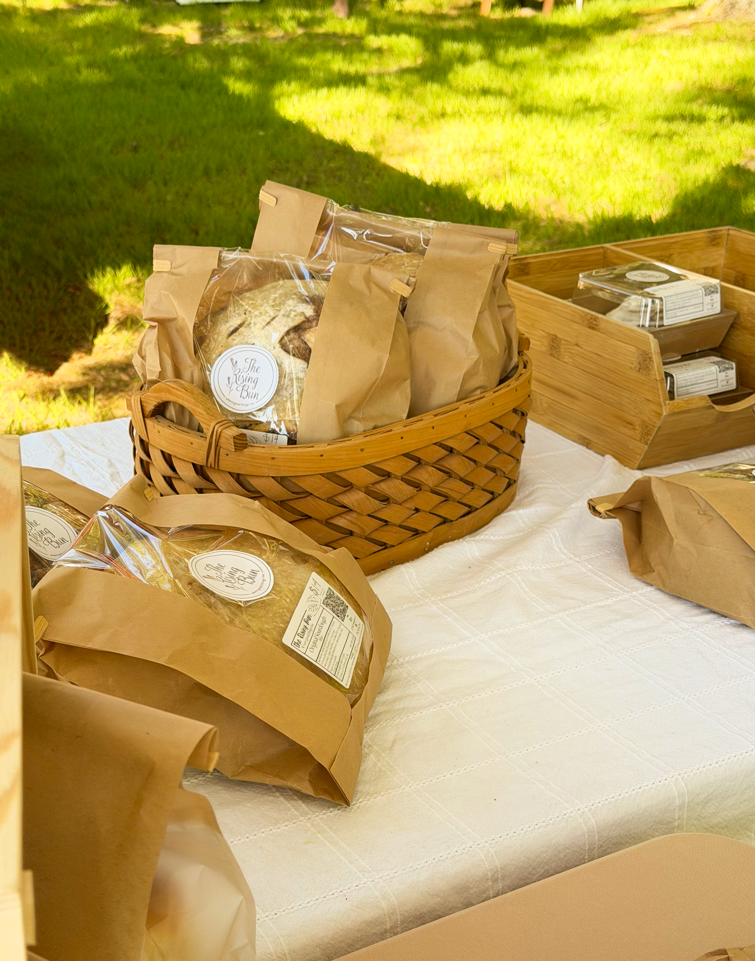 Multiple brown paper bags with clear plastic windows and food labels inside a woven basket and a wooden box on a white tablecloth outdoors on green grass.
