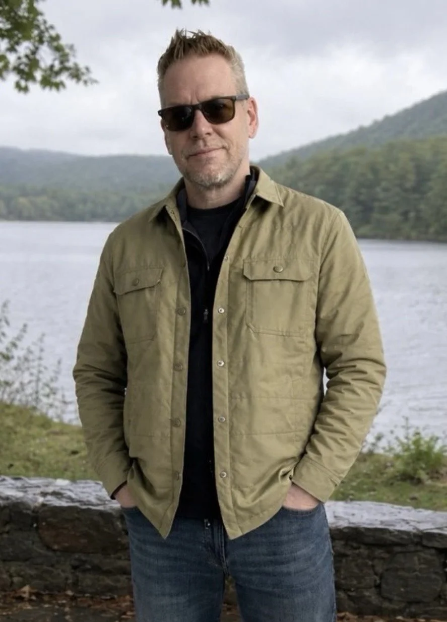 A man wearing dark sunglasses and a khaki jacket standing outdoors near a lake with trees and mountains in the background.