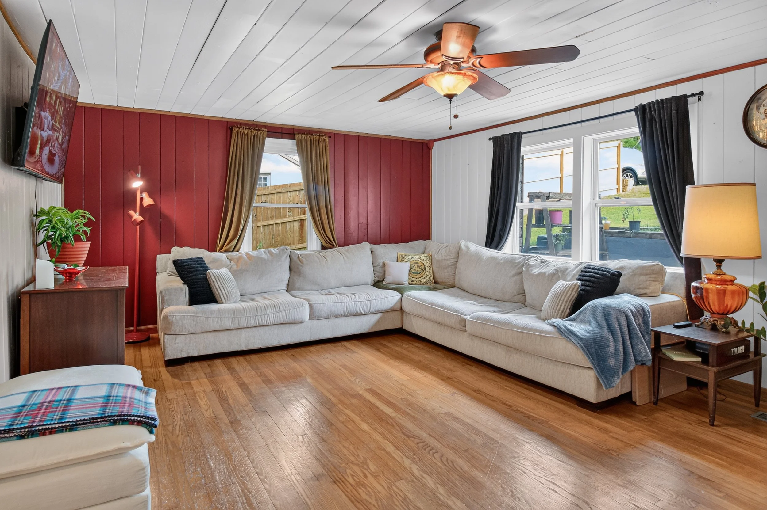 living room with white walls and ceiling, back wall painted red, beige couch, windows with curtains, orange lamp, tv, and ceiling fan.