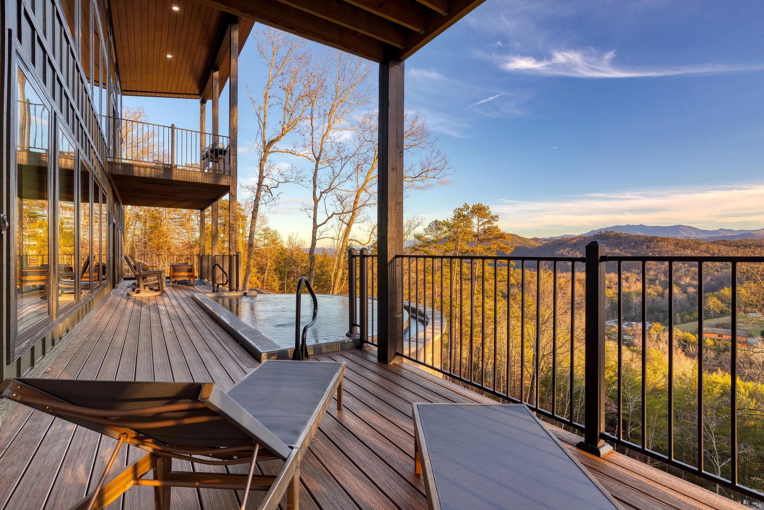 Wooden deck with lounge chairs and a small infinity pool, overlooking a valley with trees and mountains under a blue sky.